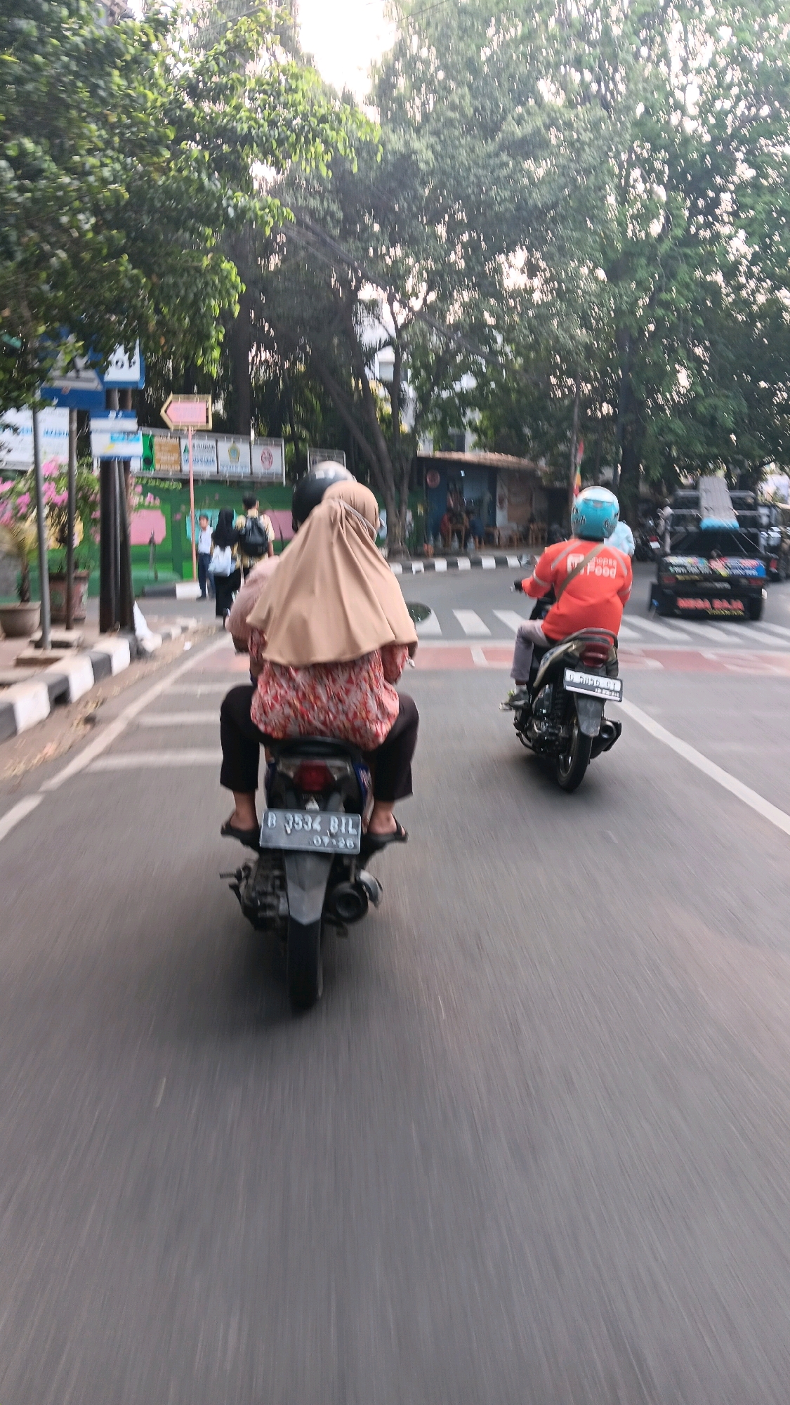 Two individuals are riding on a single motorcycle down a paved street in South Jakarta, Indonesia. The person in the rear, wearing a tan hijab and a patterned long-sleeved shirt, is likely a passenger. The person in the front, wearing an orange shirt with the text "Go Food" and a light blue helmet, is the driver. The motorcycle's license plate reads "B 3534 BTL". Trees line the street, and there are buildings and other vehicles visible in the background. The image appears to be taken in motion, indicated by the motion blur.