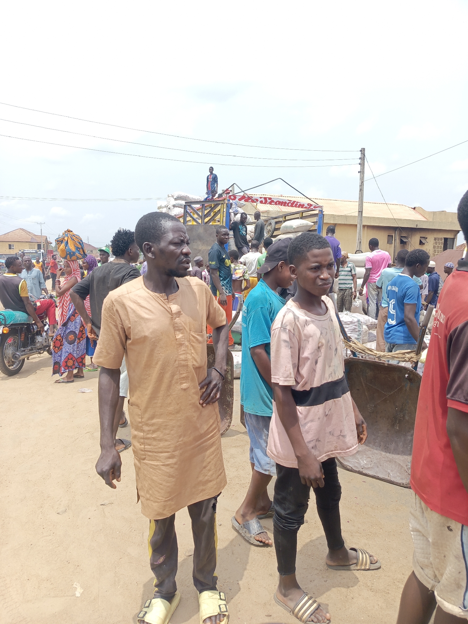 The image depicts a bustling scene in Akwanga, Nigeria. Two young men stand in the foreground on a dirt road. The man on the left wears a brown tunic over black leggings and yellow sandals. The boy to his right is in a pink and black shirt with black pants and striped sandals. Behind them, a large flatbed truck is loaded with sacks, likely of agricultural goods. People are crowded around the truck, indicating buying and selling activity. There is a man standing on the sacks that are on the flatbed truck. There is a motorcycle parked nearby, and other buildings and structures are visible in the background. Overhead power lines stretch across the sky. The lighting suggests a bright, sunny day.