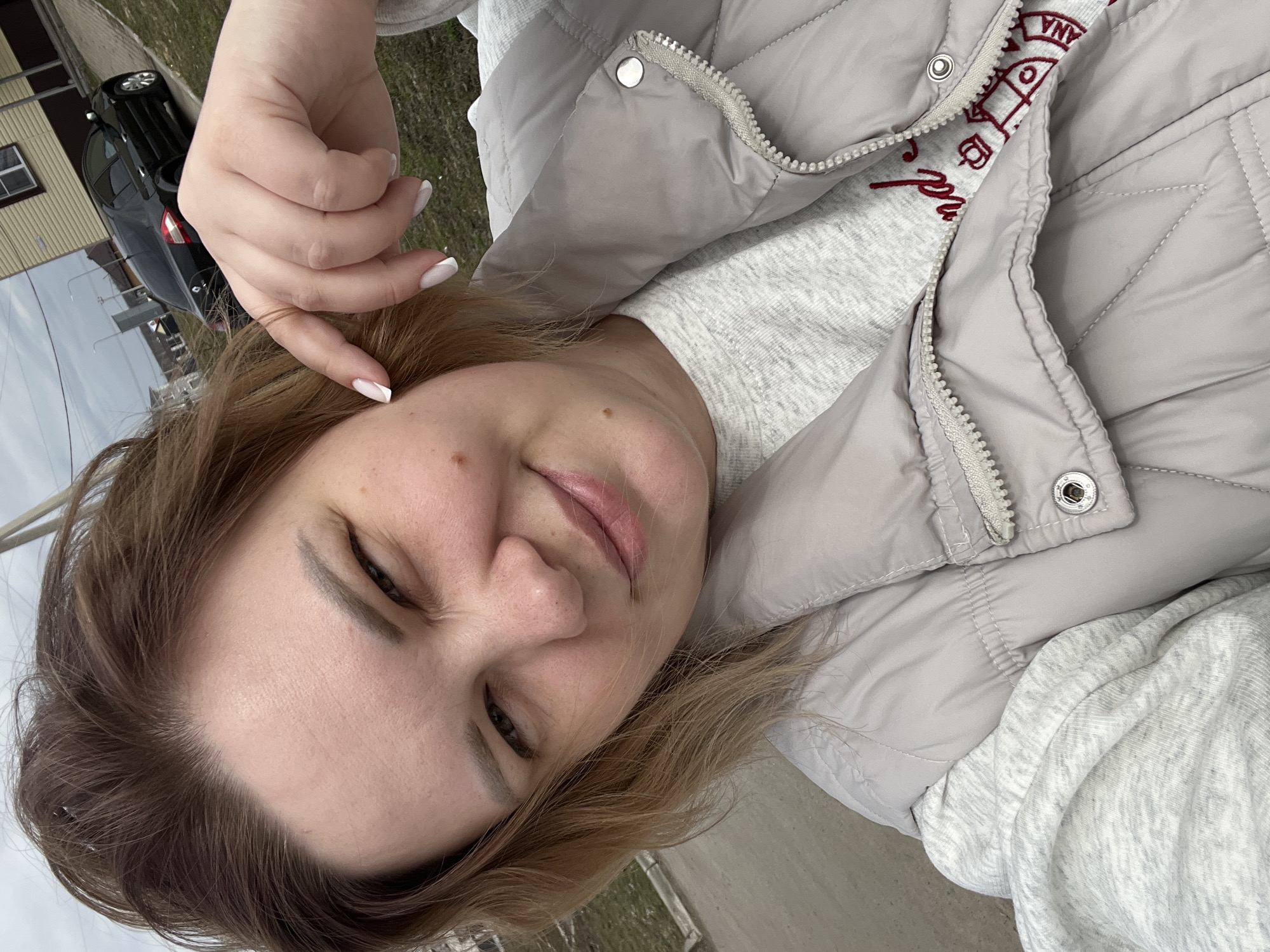 A selfie shows a woman with light skin, light brown hair, and light eyes. She is wearing a light gray quilted jacket and a light gray sweater. Her nails are manicured with a white French tip. The background includes a black car, a building with tan siding, grass, and a gray sidewalk. The location is Usman, Russia.