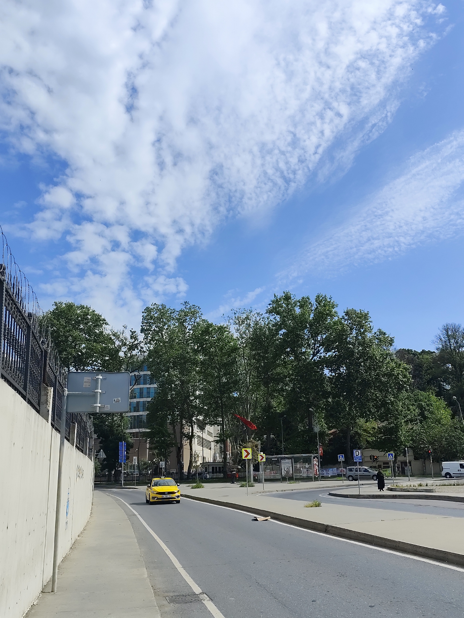 The image is a street scene in Sarıyer, Turkey, captured on a sunny day with a blue sky filled with scattered clouds. A yellow taxi is driving on the right-hand side of a gray asphalt road, marked with a white lane divider. A concrete wall with a metal fence runs along the left side of the road. Trees line the sidewalk on the right. In the distance, there is a pedestrian crossing with a woman dressed in black walking across it. A white van is parked further away. Traffic signs are also visible near the pedestrian area.