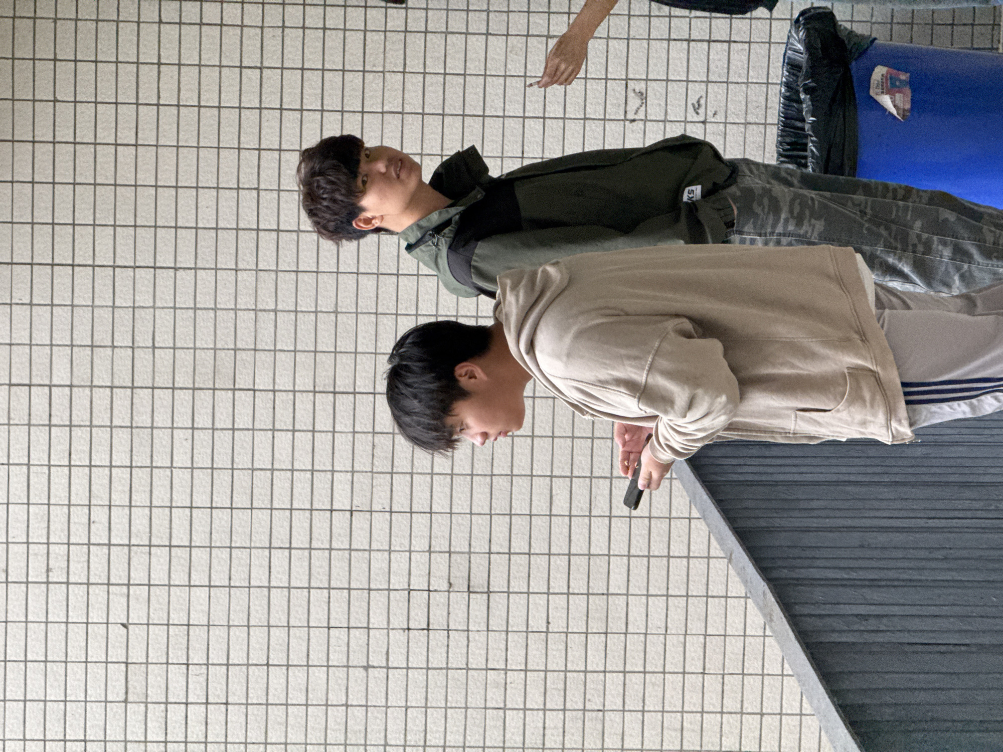 Two young men are depicted standing in what appears to be an outdoor or semi-outdoor public area with a tiled floor in Jiangjin, China. The image is oriented vertically, but described as if the subjects are standing upright.
In the foreground, a young man wearing a light beige or tan hooded sweatshirt and grey athletic pants with blue and white stripes down the side is looking downwards, holding a dark smartphone in his right hand. Behind him and slightly to his left stands another young man, dressed in a dark green and black jacket over a dark top, paired with camouflage-patterned trousers. He is looking forward and slightly upwards. The letters "QS" are visible on his jacket, likely part of a brand logo.
In the upper right background, a hand belonging to a third person is visible, holding what appears to be a lit cigarette. Adjacent to this, a blue cylindrical trash bin, partially obscured, can be seen with a black plastic bag liner protruding from it. The ground is covered with light-colored square tiles, and in the lower right, a dark, textured surface, possibly a low wall or bench, cuts diagonally across the frame. The scene appears to be set during the daytime, with soft, diffused lighting suggesting either an overcast sky or a sheltered location. The overall atmosphere is casual. No other discernible text is visible besides the logo on the jacket and an illegible label on the trash bin.