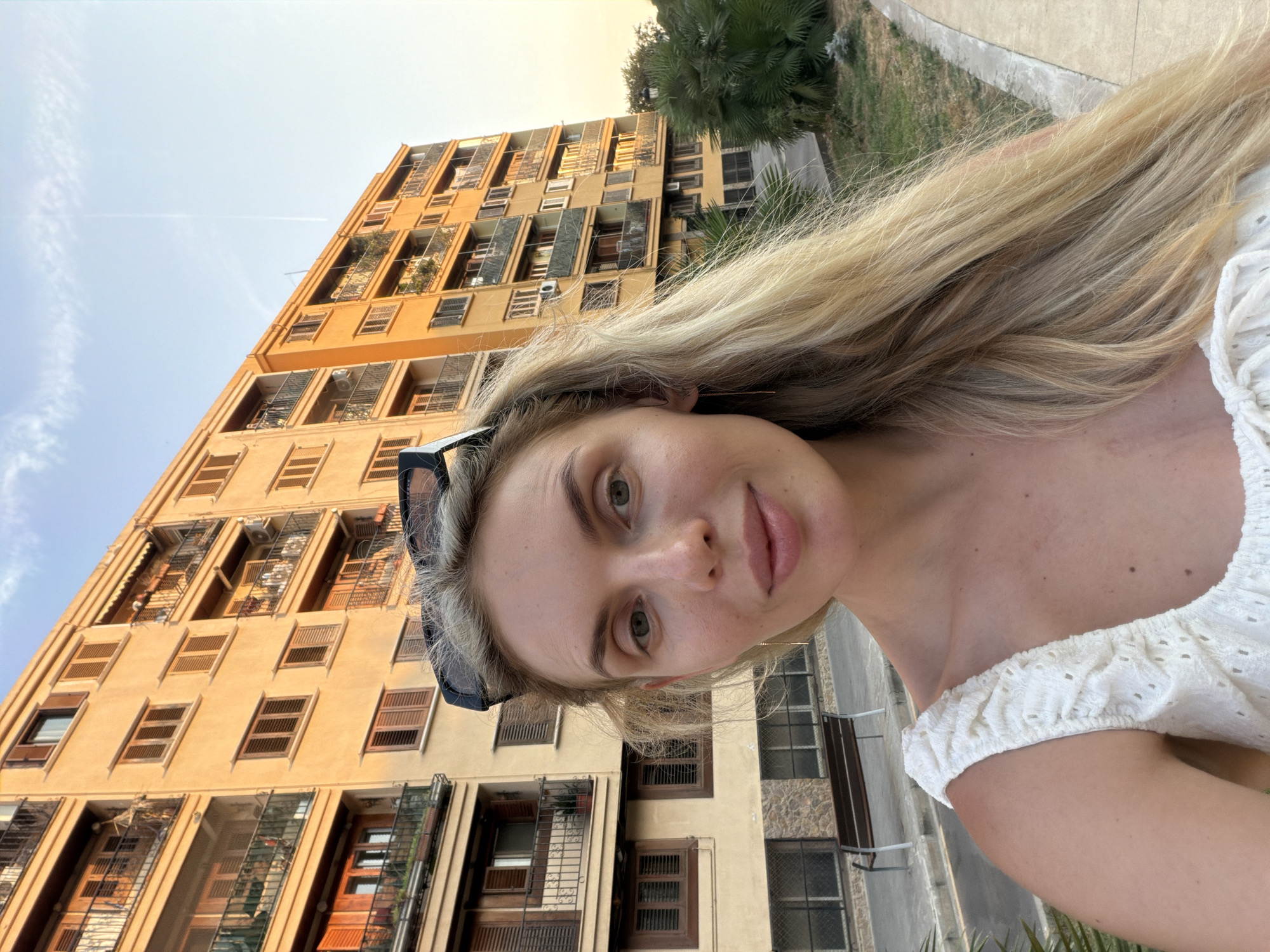 A woman with blonde hair is taking a selfie outdoors. She is wearing a white top and sunglasses are perched on her head. Behind her, a building with multiple balconies and windows is visible, bathed in warm sunlight. A paved walkway and some greenery are also present in the background. The scene appears to be in a city, possibly Benimàmet-beniferri, Spain, given the architectural style and the sunny weather.
