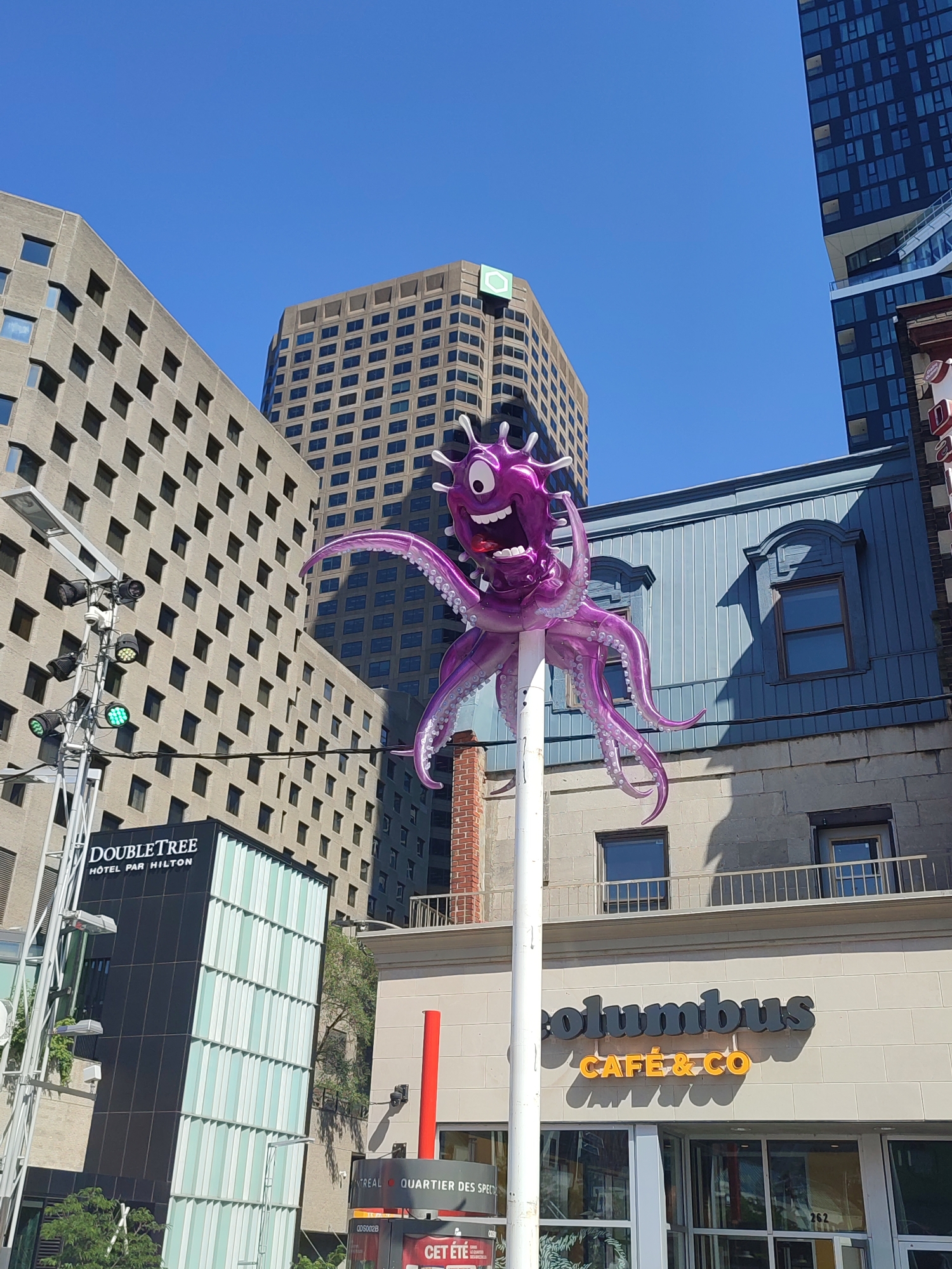 The image shows a bright, sunny day in Montreal, Canada, with a clear blue sky. A large, inflatable purple octopus sculpture is prominently displayed, mounted on a white pole. The octopus has a cartoonish face with large eyes, a wide open mouth with visible teeth, and appears to be smiling or shouting. Its tentacles are spread out.
In the background, several buildings of varying architectural styles are visible. To the left, a modern building with a black facade and a patterned glass front displays a "DoubleTree HÔTEL PAR HILTON" sign. Behind it, a taller, light brown building with many small, dark windows rises into the sky. To the right, a building with a blue clapboard facade and dormer windows is partially visible. Further to the right, a modern dark blue skyscraper with a distinctive angular design is present.
At the street level, a sign for "Columbus CAFÉ & CO" with orange lettering is visible above a storefront. A red pole and part of a street sign with French text, including "QUARTIER DES SP...", are also in the foreground. Traffic lights with green lights illuminated are mounted on a pole to the left of the DoubleTree building. The overall scene suggests an urban environment with a mix of businesses and modern and older architecture. There are no people visible in the image.