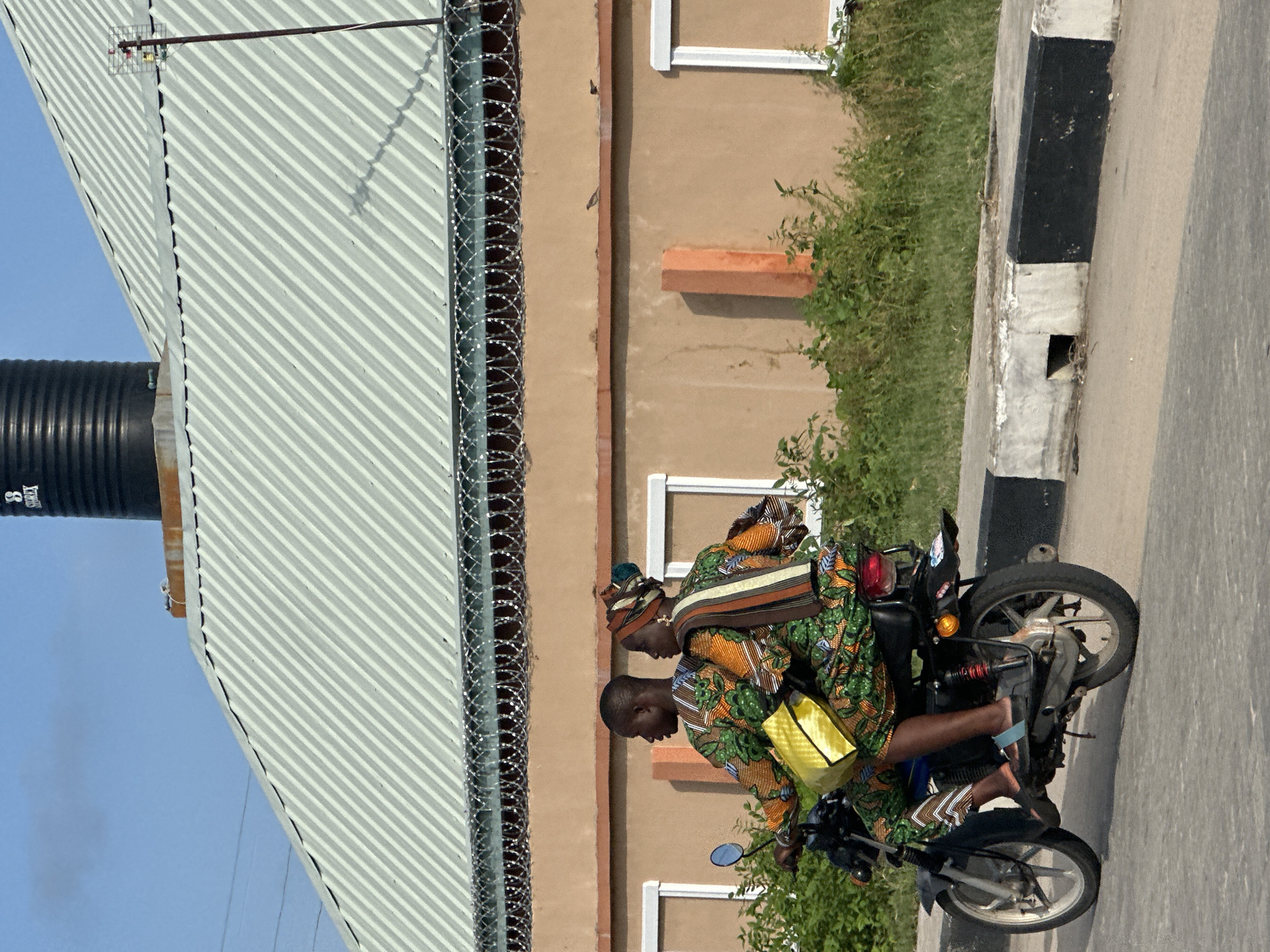 Two people are riding a motorcycle in an outdoor scene. They are both wearing colorful patterned clothing, with the person in the back also wearing a yellow bag. The motorcycle is black with some red accents. The setting appears to be a street with a building in the background that has a corrugated metal roof. There is also some greenery and a concrete curb. The image is taken during the day, and the weather appears to be clear. There is no visible text in the image. The location is Badagry, Nigeria.