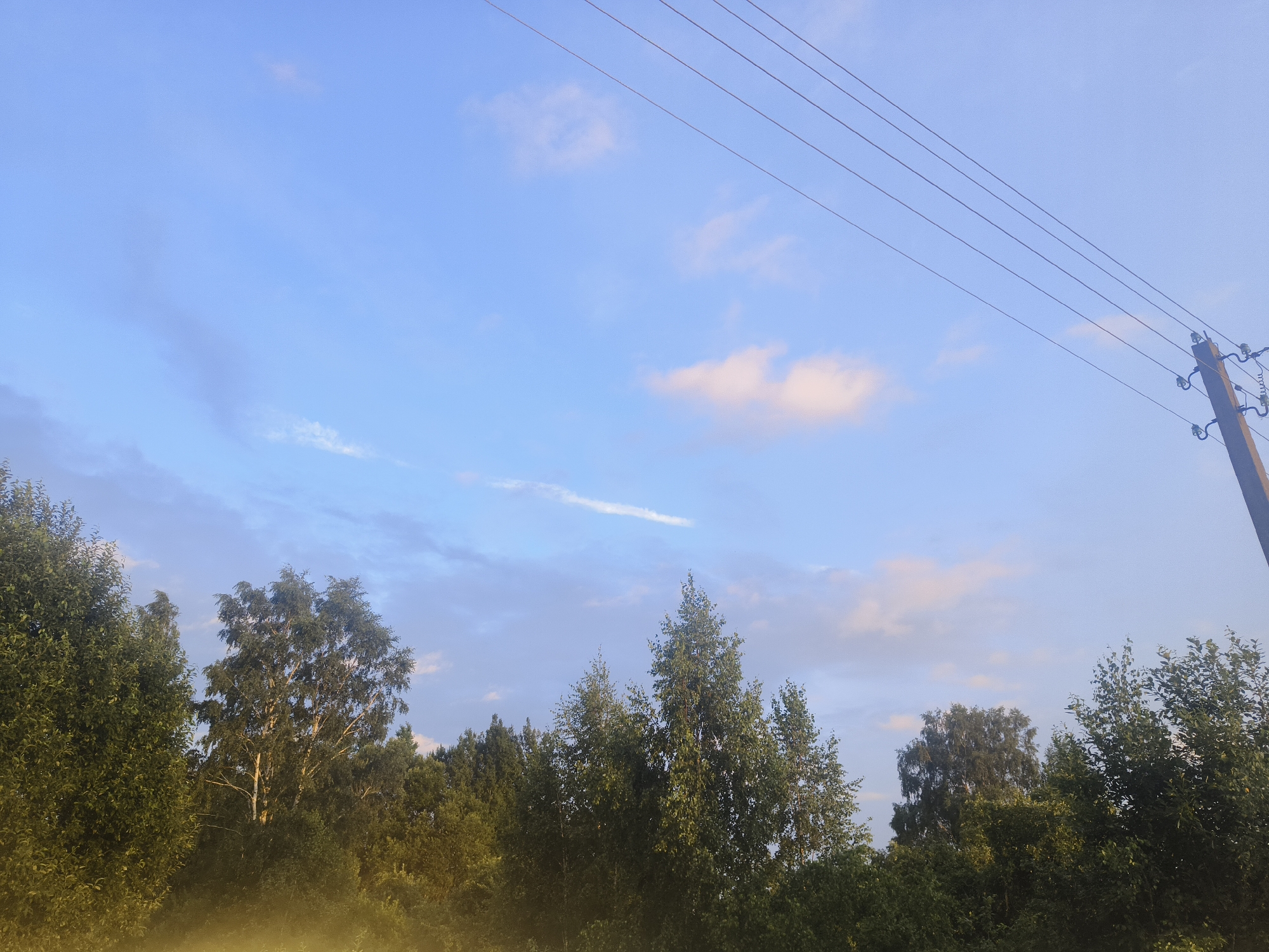 The image captures an outdoor scene with a clear blue sky dotted with a few wispy clouds. Several parallel power lines stretch diagonally across the upper right portion of the frame, connected to a utility pole on the far right. Below the sky, a dense line of green trees forms the horizon, with varying heights and leaf densities. The foliage appears lush and suggests a natural, possibly rural environment. In the foreground, a hazy, yellowish mist or light filters upwards from the bottom edge, obscuring the lower parts of the trees. The overall impression is a tranquil, natural setting under a clear sky, possibly during early morning or late afternoon given the quality of light. The location is identified as Kruglitski sielski Savek, Belarus. There are no people or animals visible in the image.
