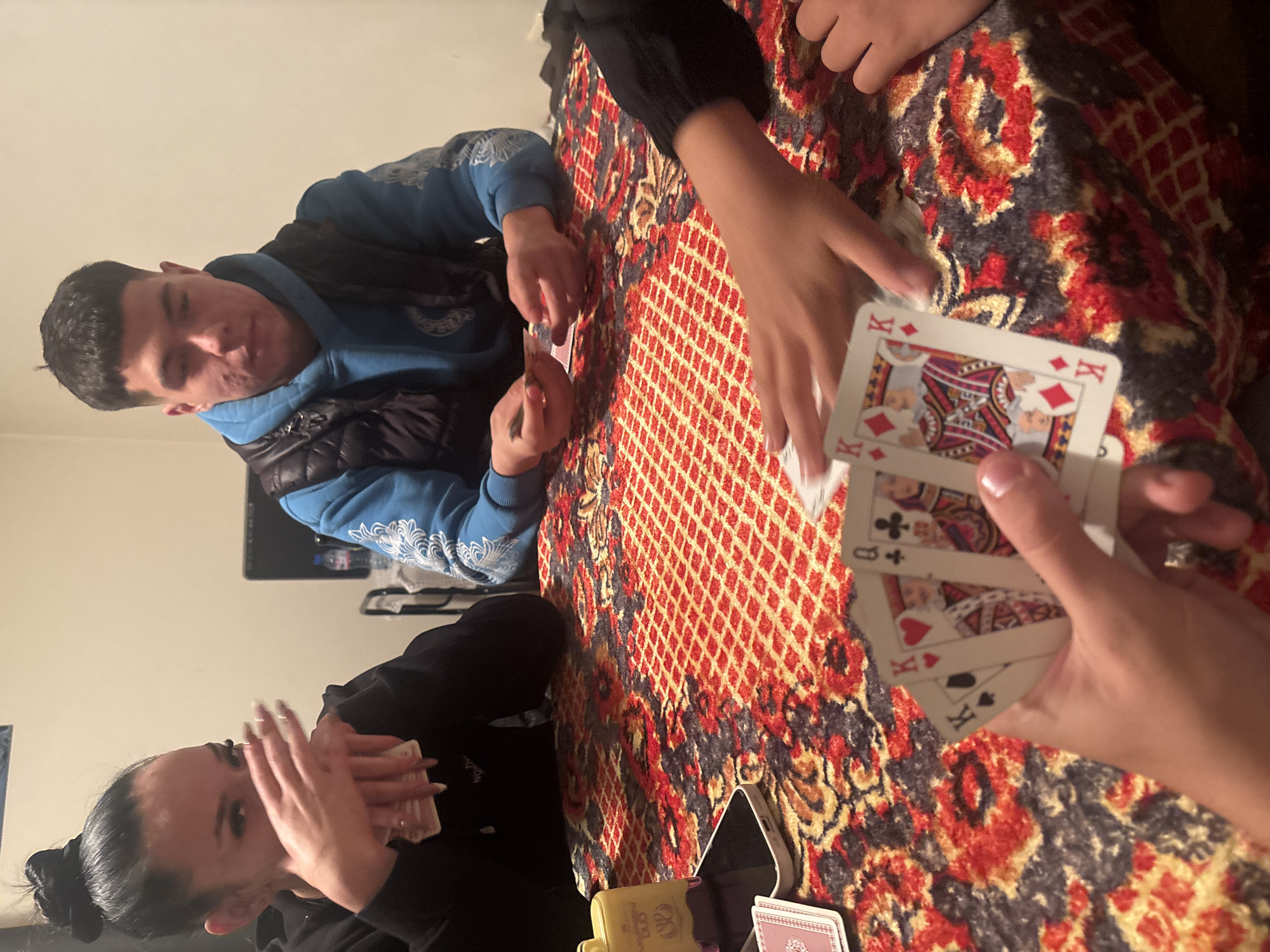 Three people are sitting around a table with a patterned red and brown tablecloth, playing cards. One person, in the lower left, has dark hair pulled back and is holding cards close to their face. Another person, on the left side of the table, is wearing a blue hooded sweatshirt and a black vest, and also holding cards. A hand holding cards is in the foreground. The cards visible are a King of Diamonds, Queen of Clubs, King of Hearts, and King of Spades. A yellow card box and a cell phone are on the table near the bottom of the image. A television is visible in the background. The location is not identifiable as Goritsa, Bulgaria based solely on this image.