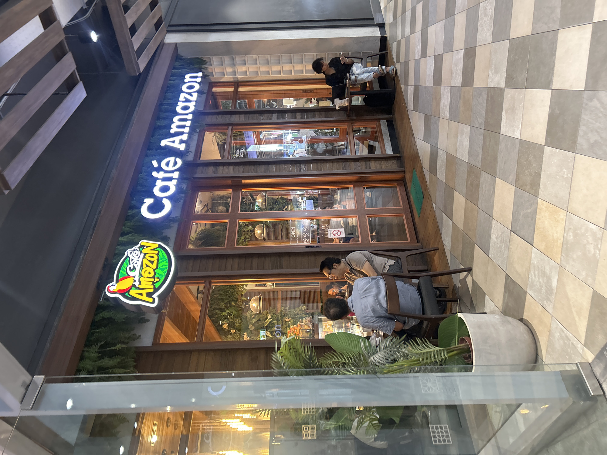 The image shows the exterior of a "Cafe Amazon" located in Bangkok, Thailand. The cafe has a distinctive wooden facade with large glass windows. A prominent neon sign with a parrot logo and "Cafe Amazon" is visible above the entrance. Inside, two men are seated at a table, facing away from the camera, appearing to be engaged in conversation. Another person is seated at a small table outside the cafe, looking at their phone. The floor is tiled in a checkerboard pattern of beige and grey squares. Green plants are visible both inside and outside the cafe, adding to the natural aesthetic. The overall environment suggests a modern, somewhat upscale cafe setting within an urban area.
