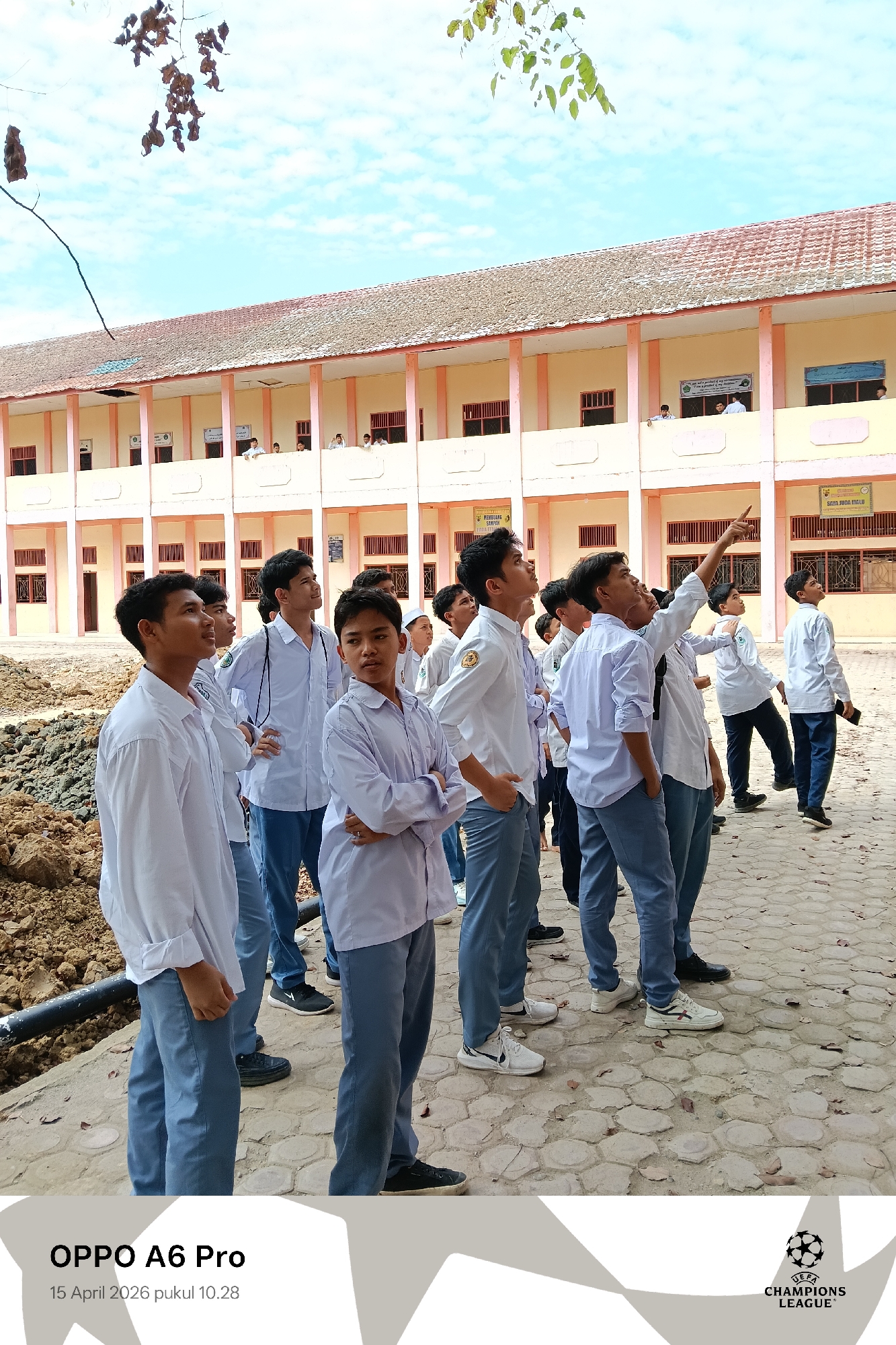 The image depicts a group of approximately ten young men, likely students, standing outdoors and looking upwards. They are dressed in similar attire: white shirts and grey trousers, suggesting they are in school uniforms. One student has his arms crossed.
Behind them is a two-story building with a red-tiled roof and a light-colored facade. There are visible windows and some signage on the building. A construction site with exposed earth and pipes is present to the left of the group.
The ground appears to be a paved area with a pattern of hexagonal tiles. The sky is visible in the background, indicating it is daytime.
The image includes text at the bottom indicating it was taken with an OPPO A6 Pro on 15 April 2026 at 10:28. The UEFA Champions League logo is also present. Based on the visible signage in the background (SAYA JUDA MALU), it is likely in the city of Lhokseumawe, Indonesia.