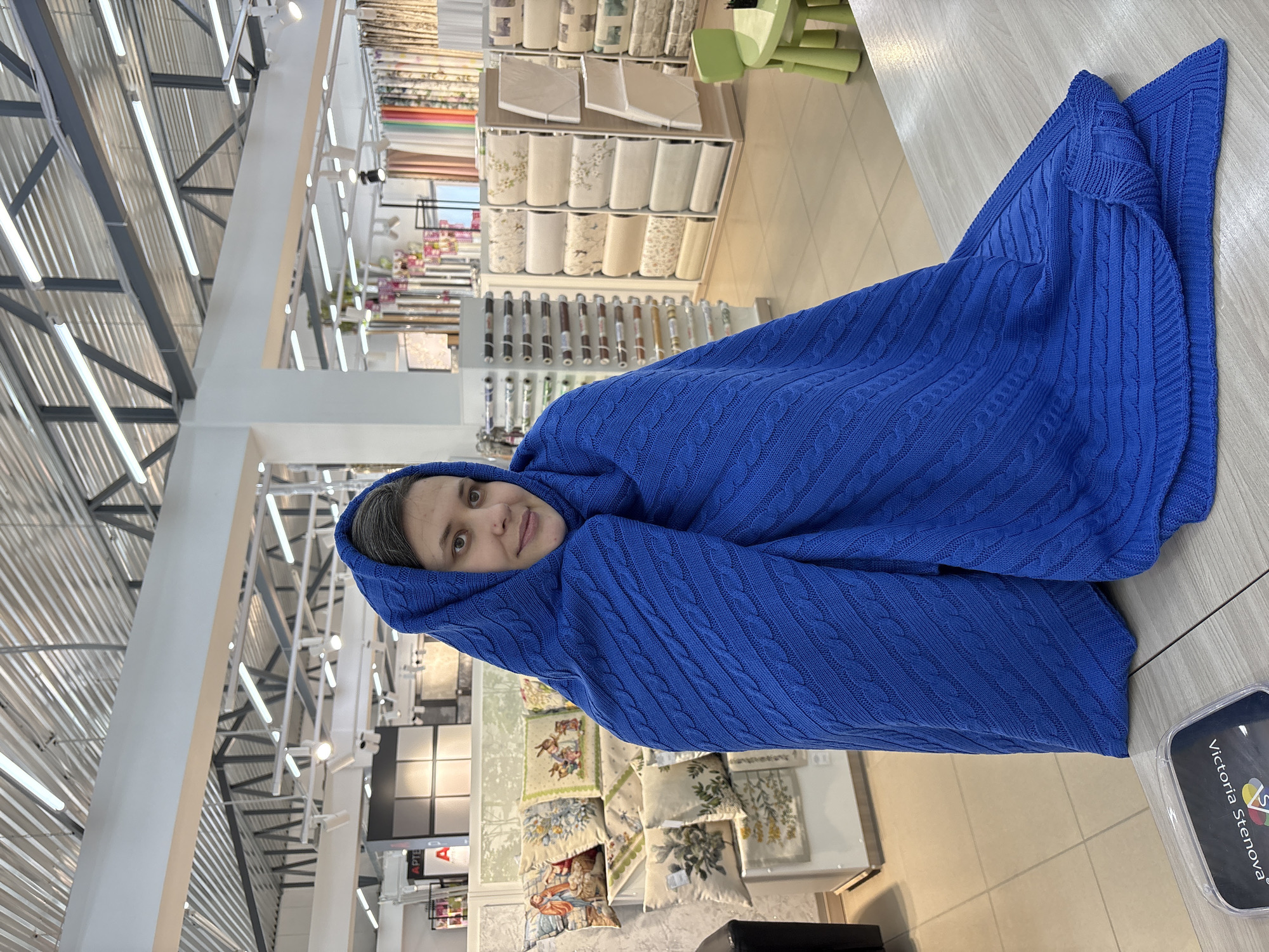 A vertical eye-level shot depicts a person wrapped in a blue cable-knit blanket, positioned in an interior space that appears to be a retail store. The person, likely a woman, has medium-length brown hair and is looking directly at the camera with a slight smile. The blue blanket drapes over her head and shoulders, obscuring her body.
In the background, shelves display various types of fabrics, linens, and other home goods, suggesting the store specializes in textiles or interior design. Lighting fixtures are visible on the ceiling, indicating a modern commercial setting. The city of Chaikovsky, Russia, is only available in the image name and is not explicitly depicted in the scene.
The photo's composition and the subject's pose give the impression of someone playfully interacting with the store's merchandise. A plastic tag with the text "Victoria Stenova" is visible on the right side of the image.