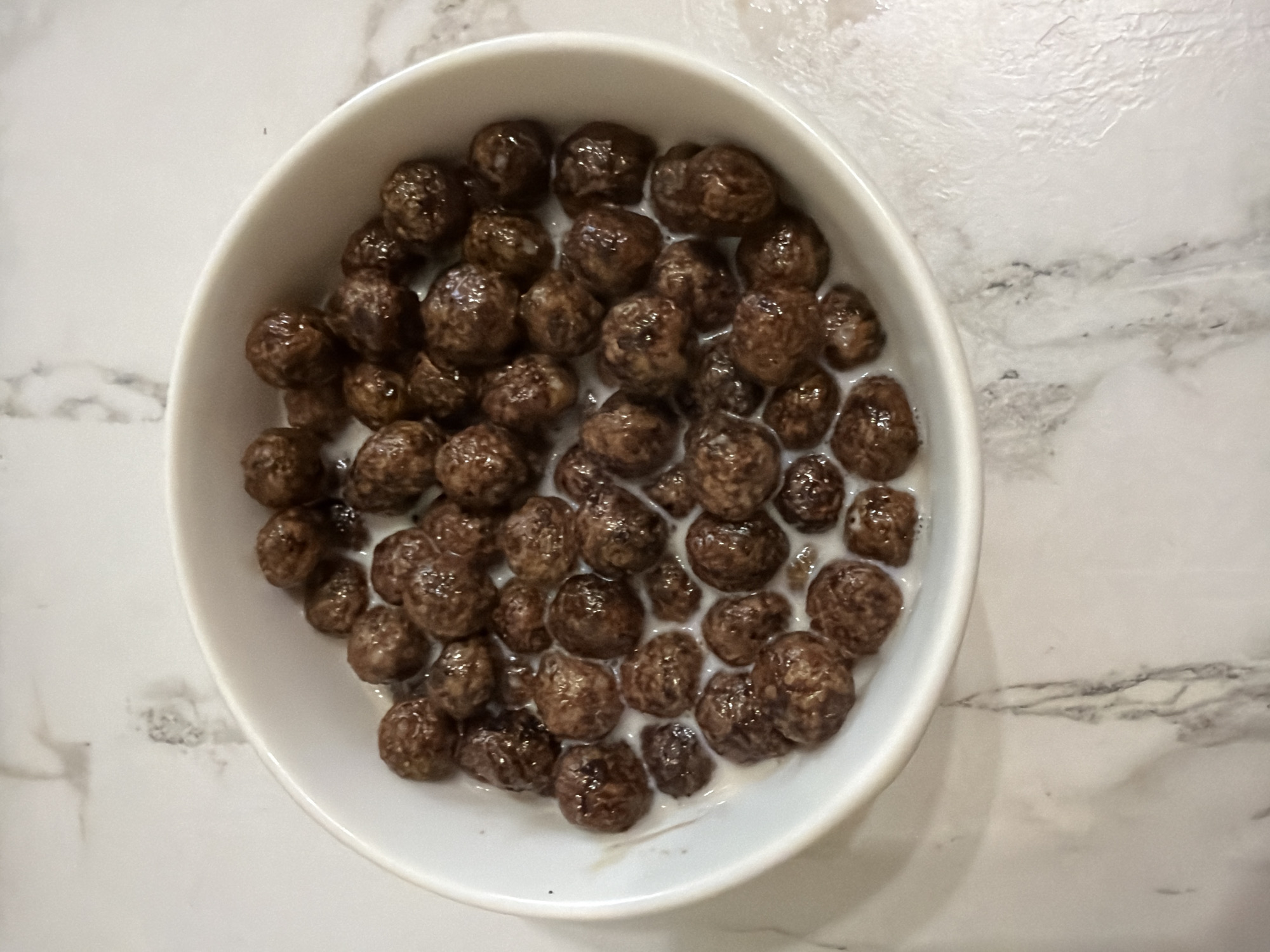 A white bowl filled with chocolate cereal and milk sits on a marble countertop. The cereal pieces are round and appear to be coated in chocolate. The milk is partially submerged the cereal.