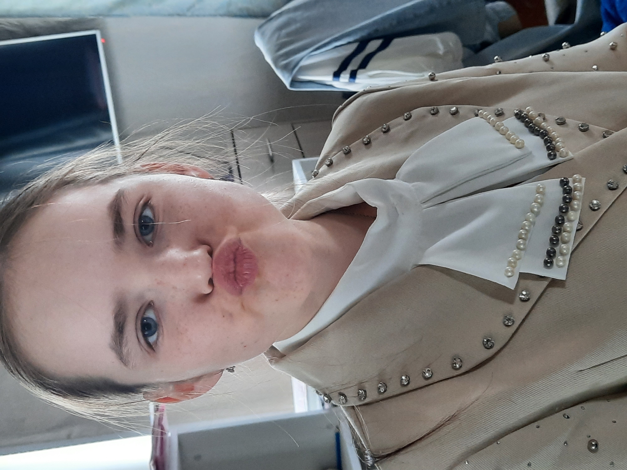 The image shows a fair-skinned person with blue eyes and light brown hair looking at the camera with pursed lips. The person is wearing a beige jacket with a white bow. The jacket is decorated with pearls and rhinestones. A television is visible in the background. The location is not discernable.