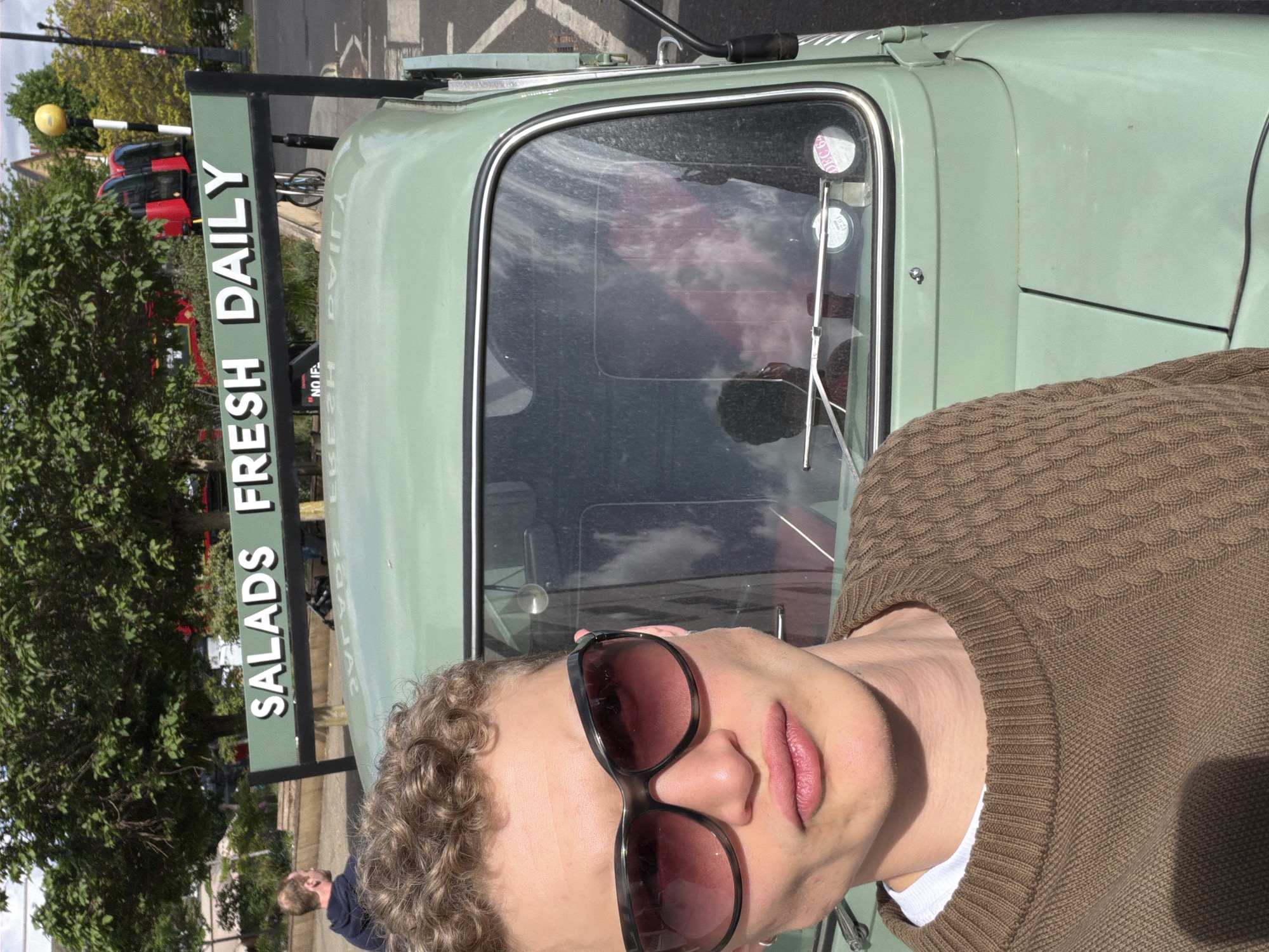 A person with curly hair and sunglasses stands in front of a light green food truck. The truck has a sign that reads "SALADS FRESH DAILY." The background includes trees, a street with cars (including red double-decker buses), and buildings. The location is likely in London, United Kingdom, based on the presence of the double-decker buses and the style of the buildings.