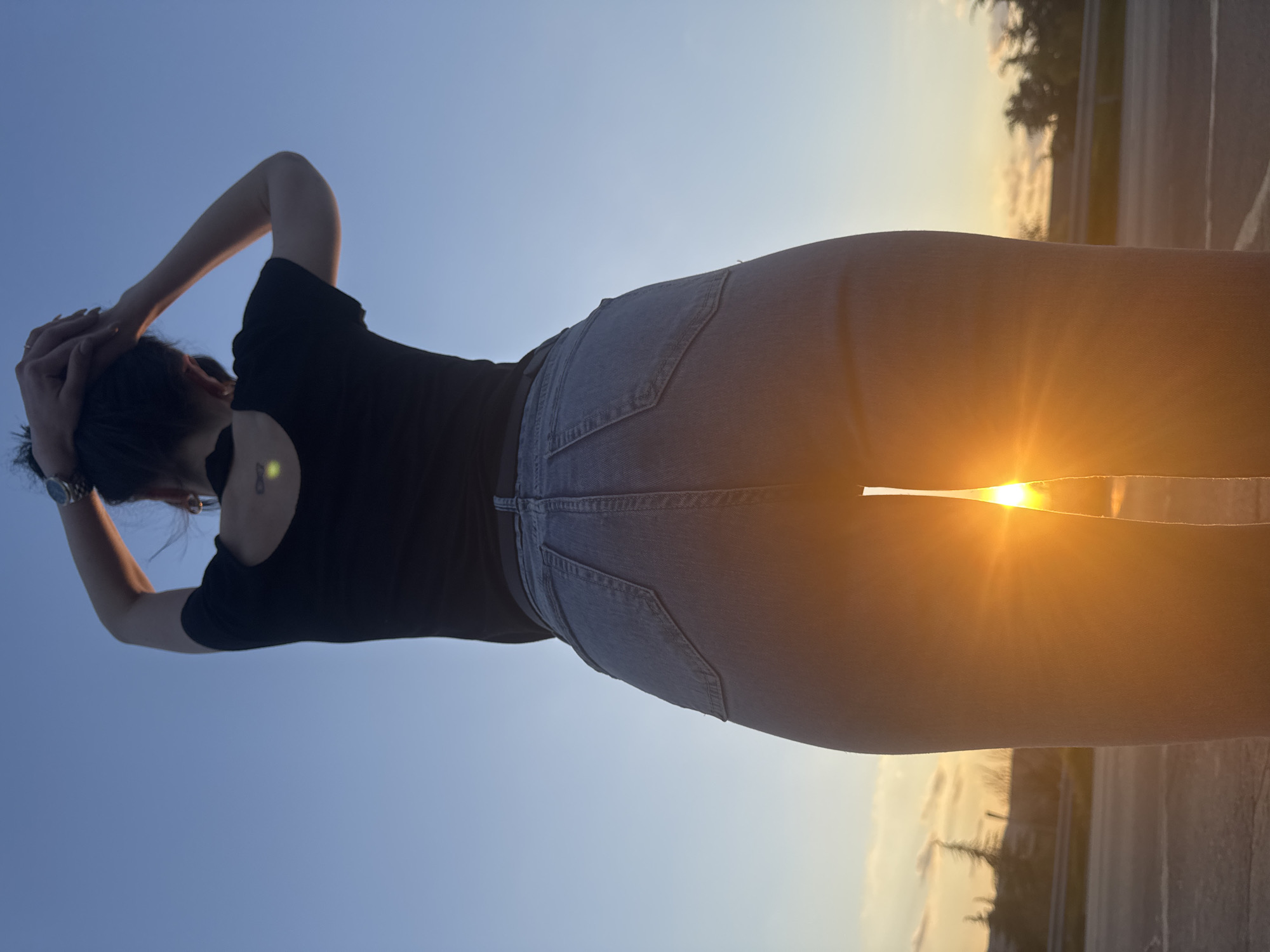 A woman stands outdoors against a backdrop of a clear, blue sky transitioning to a golden sunset. She is facing away from the viewer with her hands raised and clasped behind her head. She is wearing a short-sleeved black shirt and light blue denim jeans. The sun is directly behind her lower back, creating a bright, golden glow. The scene appears to be alongside a road or highway, with a blurred background of trees and what appears to be a barrier or curb. There is no indication of this photo being taken in Kipra, Bulgaria, or anywhere else.