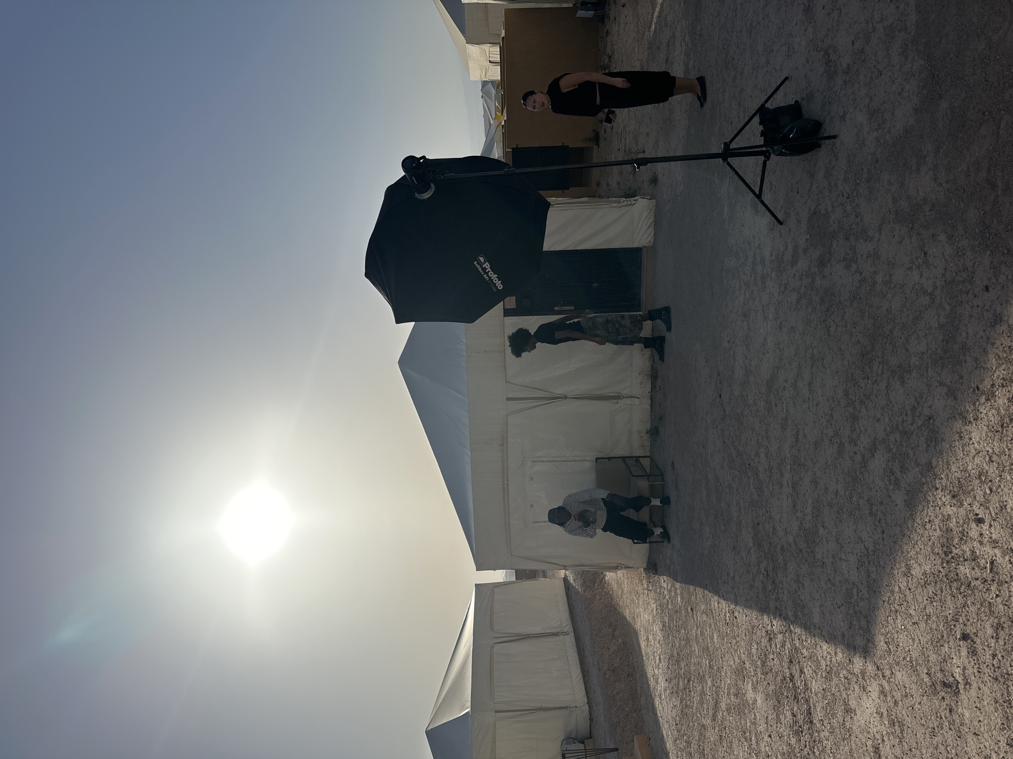 The image depicts an outdoor setting with bright sunlight. There are several people present: one standing in the background dressed in all black, another standing near a white tent, and a third person sitting on a chair in front of a tent. The scene includes white tents, indicating a possible event or encampment. A large black Profoto softbox is prominently positioned on a stand. The location is Sidi Badhaj, Morocco, suggested by the terrain and architectural style.