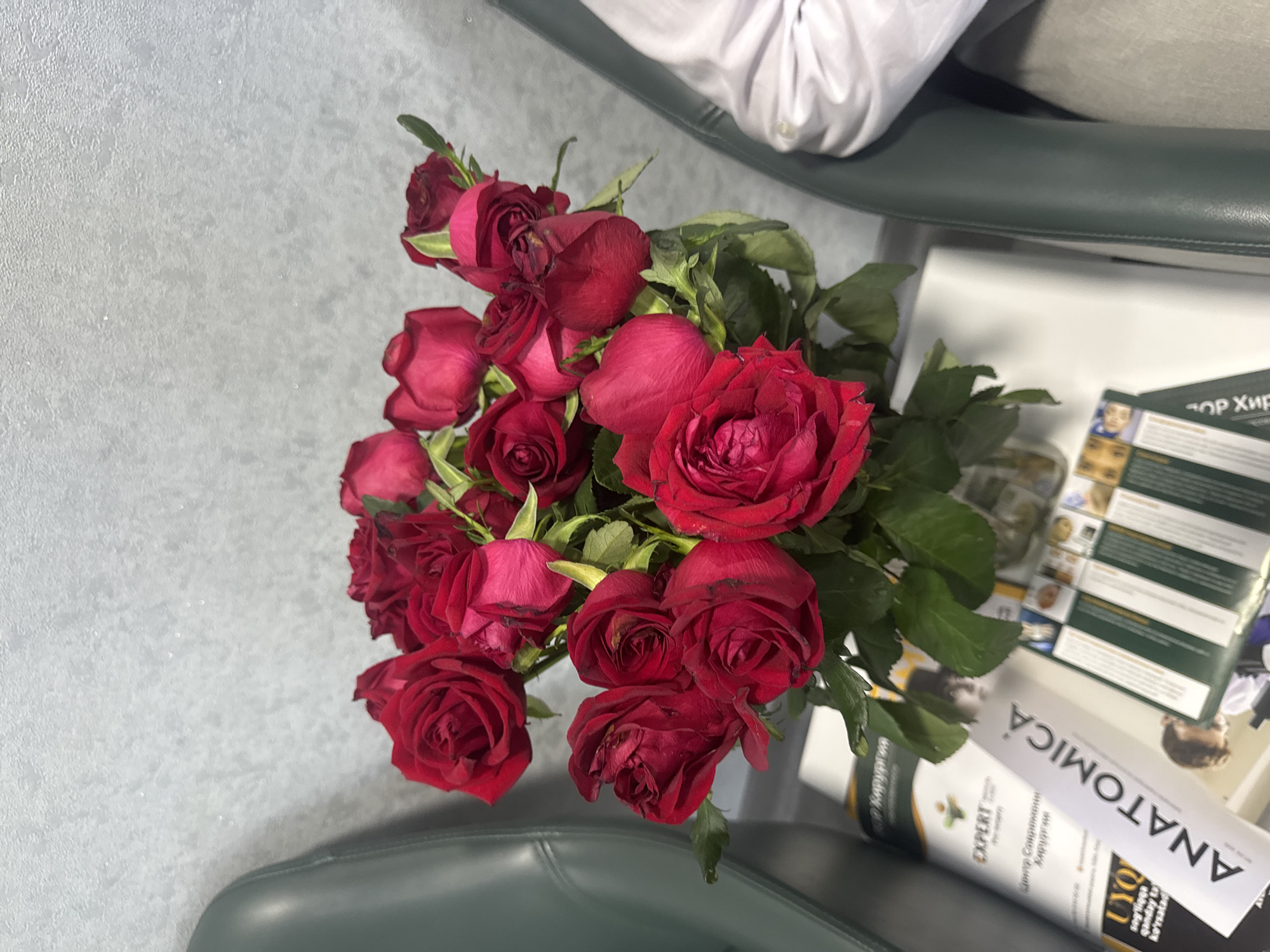A bouquet of red roses is arranged in a glass vase, placed on a white surface next to some magazines. The background features a textured gray wall and parts of two dark green chairs. One magazine on the left has the title "ANATOMICA" in large letters. The scene appears to be indoors, possibly in a waiting room or office. There are no visible people or ongoing actions. The lighting is even, suggesting an artificial light source. The composition is top-down, with the roses being the primary focus. The image does not contain any discernible landmarks or features that would indicate its specific location within Tashkent, Uzbekistan.