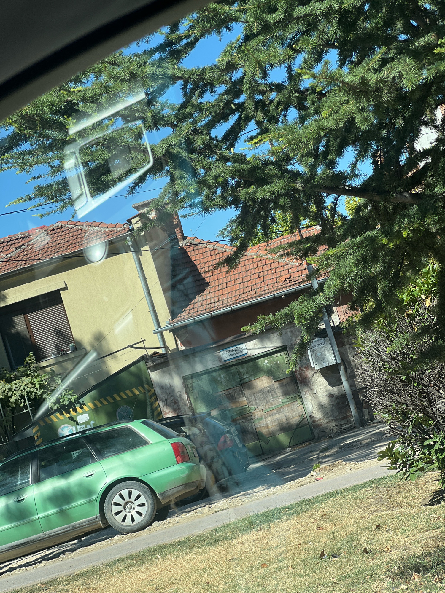 This image depicts a street scene, likely in Plovdiv, Bulgaria, viewed through a car window, with branches of a coniferous tree obscuring parts of the view. A green station wagon is parked on the left side of the frame, facing away from the viewer. Behind the car, a building with a terracotta tiled roof is visible. A garage door, painted green and weathered, is present in the middle ground. Another building, likely adjacent to the garage, is also visible. The ground is covered with dry grass and a paved surface. The sky is clear and blue. A sign is attached to the wall above the garage door, but its text is partially obscured and difficult to decipher. There are no people clearly visible in the image, though a shadow suggests the presence of a person near the garage.