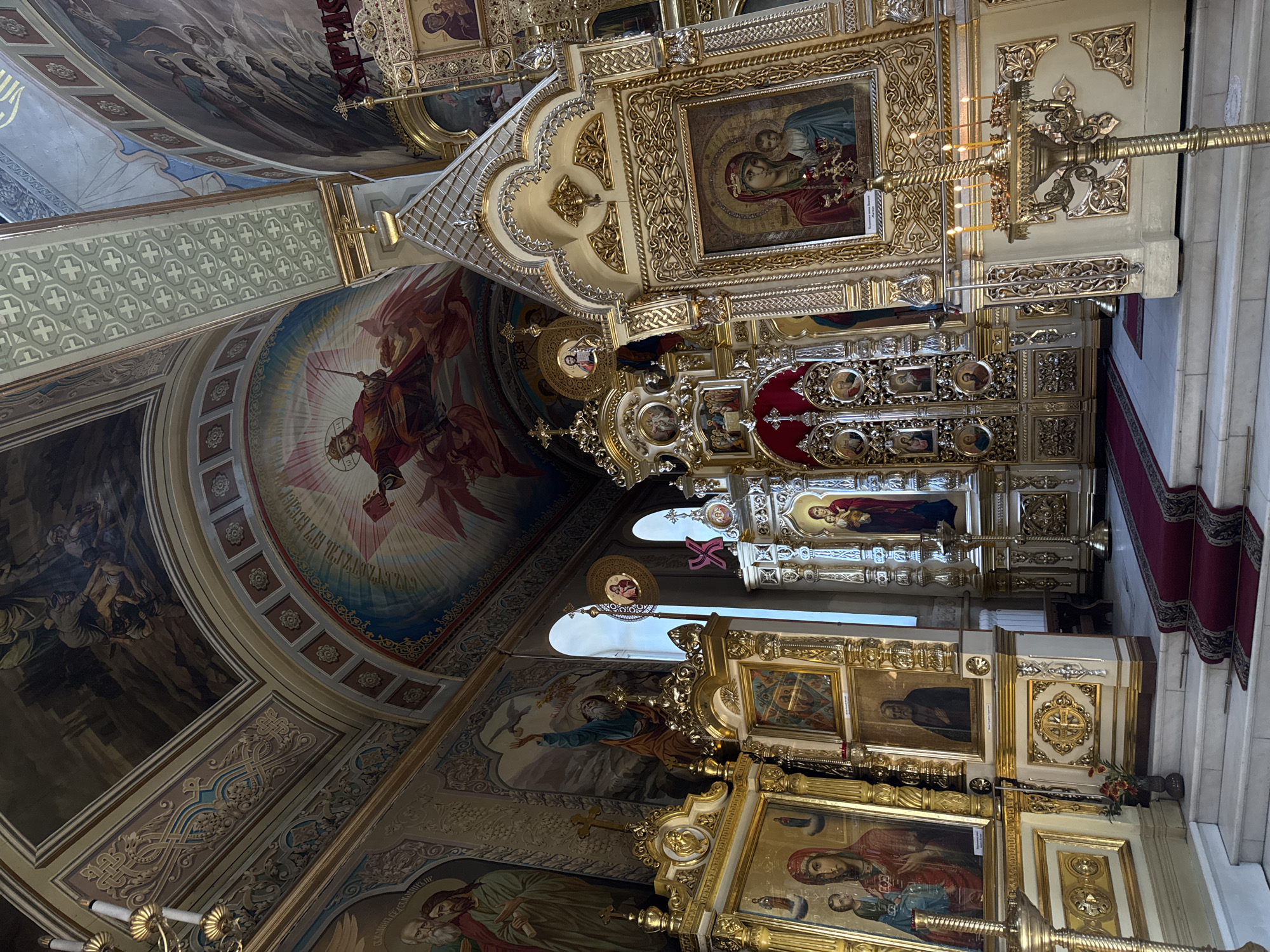 The photograph shows the interior of an ornate church, likely Orthodox Christian, with richly decorated walls and ceiling adorned with religious murals and iconography. In the foreground, a gilded iconostasis, a wall of icons and religious images, dominates the scene. The iconostasis is elaborately carved and gilded, featuring several icons, including depictions of the Virgin Mary and Jesus. Candles are lit on a golden candelabra in front of the iconostasis. A red carpet leads up a marble staircase towards the iconostasis. The ceiling is painted with scenes of biblical figures and celestial imagery, with intricate decorative moldings and gilded accents. The overall impression is one of grandeur and spiritual reverence.