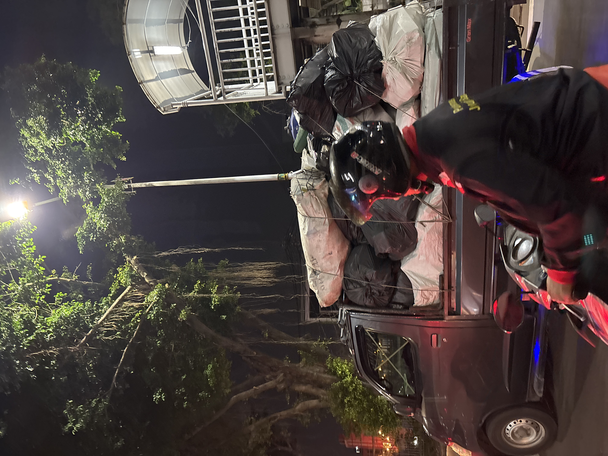 In West Jakarta, Indonesia, at night, a dark-colored pickup truck is heavily loaded with large sacks, some white and some black. The truck is parked on a street with a pedestrian overpass visible in the background, illuminated by a streetlamp and overhead lights. A person wearing a black helmet and a black jacket with yellow writing is riding a motorcycle in the foreground, passing by the truck. The motorcycle has red and blue underglow lights. The scene is dimly lit, with the primary light sources being the streetlamp and the lights on the overpass and motorcycle. A large tree with hanging roots is visible on the left side of the frame, casting shadows.