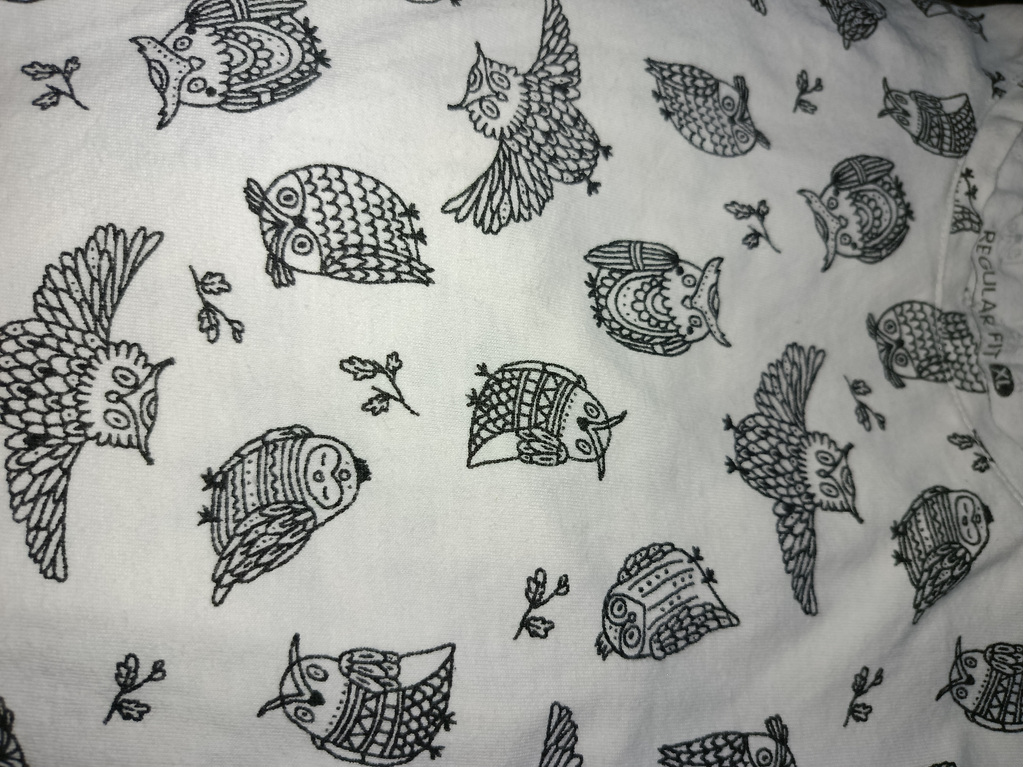 A close-up, overhead shot of a white t-shirt with a repeating pattern of black and white owls. The owls are drawn in a stylized, folk-art manner. Some owls are depicted flying, while others are perched. There are also small, stylized branch-like elements scattered between the owls. The fabric appears to be a textured cotton. A white tag with black text is visible on the right side of the image, partially obscured by the fabric, with the words "REGULAR FIT" visible. The lighting is even, suggesting an indoor environment.