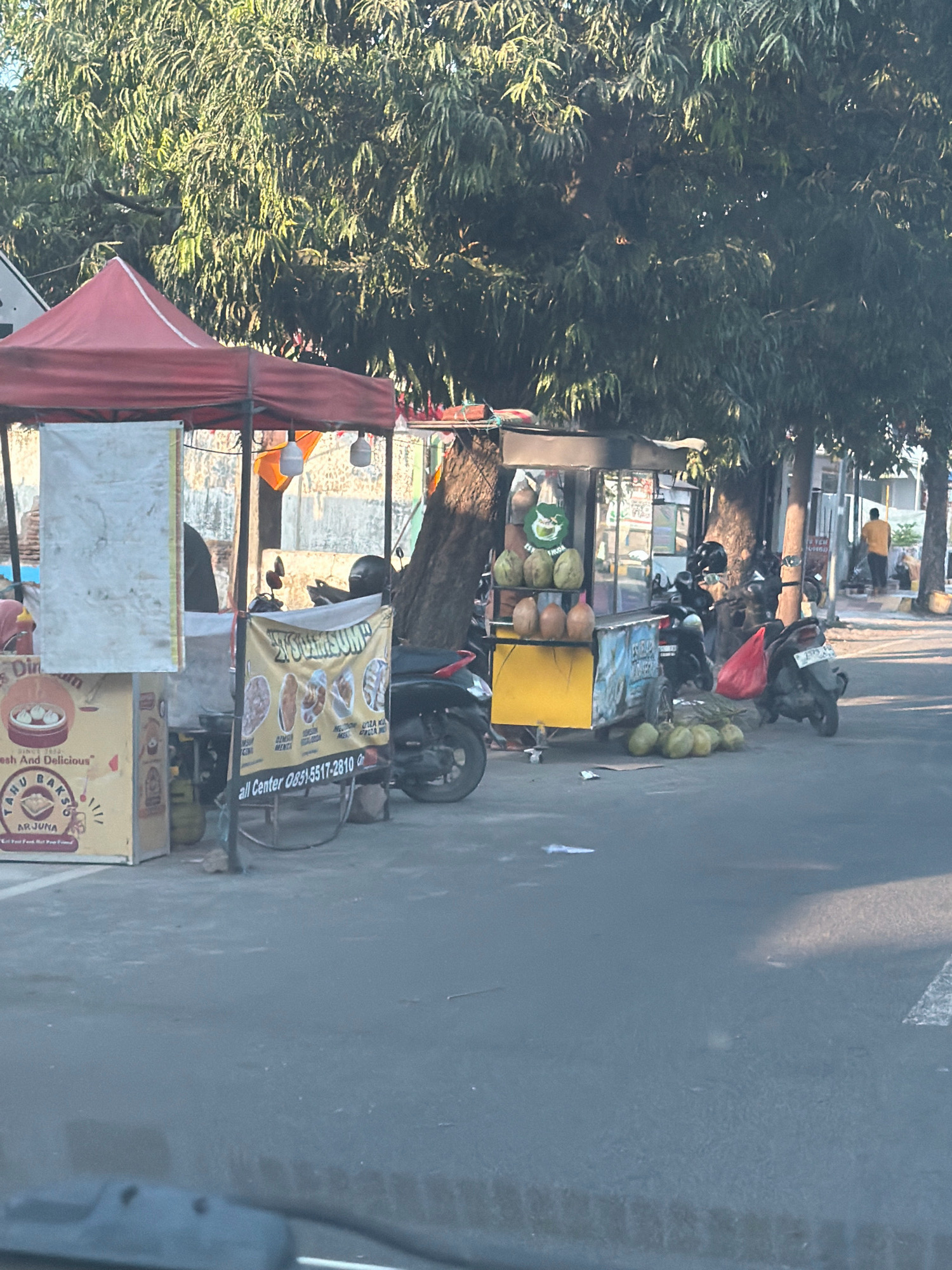 A street scene in Situbondo, Indonesia, features several food stalls set up along the side of a road. The stalls are adorned with red and orange awnings, and some are decorated with signs and banners. One stall displays a selection of coconuts, while another offers what appear to be fried snacks. Several motorcycles are parked nearby, suggesting a busy area. In the background, a person in an orange shirt walks down the street, and a building with a light-colored facade is visible. Lush green trees with dense foliage provide shade over the scene. The overall impression is of a vibrant local market area, bustling with activity and the aroma of food.