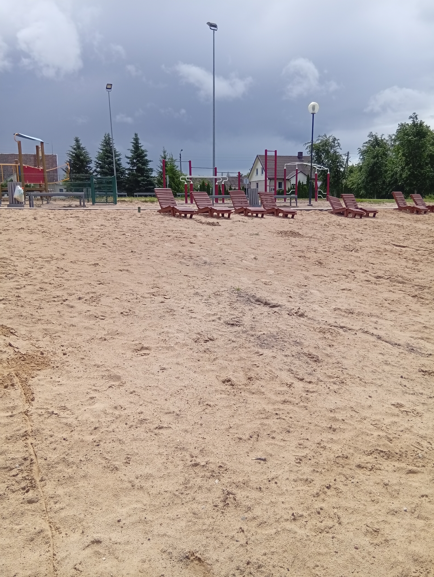 The image shows a sandy outdoor area, likely a beach or playground, in Šaukėnai, Lithuania. In the foreground, the sand is undisturbed, with small pebbles and indentations visible. In the mid-ground, several wooden lounge chairs are arranged on the sand. Behind the chairs, there is a playground structure with red and yellow elements, along with a metal fence and trees. In the background, there are residential buildings and more trees under a cloudy sky. Tall streetlights are also present. The scene depicts a public recreational space.