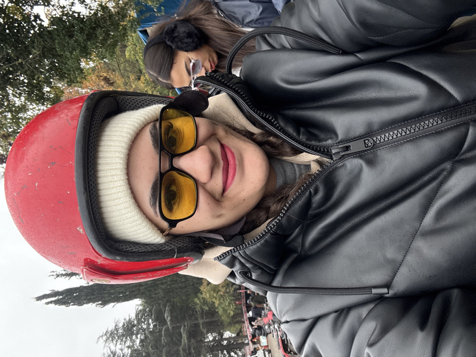 The image captures an outdoor scene in Çiftlikköy, Turkey, under an overcast sky. In the foreground, a female subject, appearing to take a selfie, is prominently featured. She wears a red full-face helmet over a white knit beanie, yellow-tinted sunglasses with a black frame, and a black faux leather puffer jacket with a visible zipper, worn over a beige layer. Her lips are covered in dark pink lipstick, and she displays a smile with visible dimples. To the right and slightly behind her, a second female is partially visible, wearing black fluffy earmuffs and light-framed, lightly tinted sunglasses, looking downwards and to the right. The background features tall, dark green coniferous trees, dense green foliage, and several all-terrain vehicles (ATVs) with individuals gathered around them, indicative of an ATV riding or rental area. Red and white striped barrier tape is discernible in the left background near the ATVs. A blue tarpaulin or structure is faintly visible on the far right side.