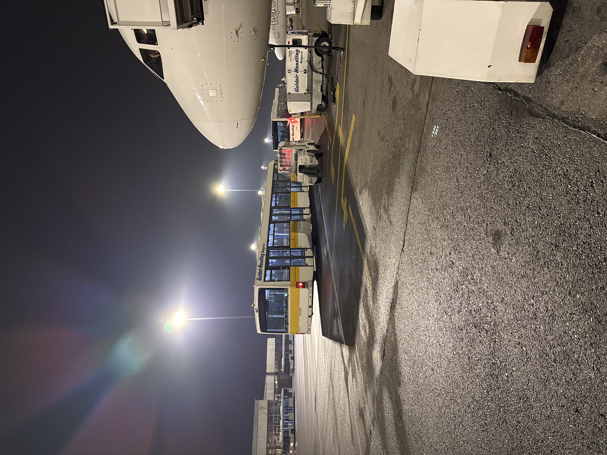 The image depicts a scene at Sofia Airport in Sofia, Bulgaria, at night. A large white airplane dominates the upper left corner, with a passenger bus parked alongside. The bus, bearing the logo of "Goldair Handling Bulgaria," is illuminated by streetlights. Airport infrastructure, including gates and additional service vehicles, is visible in the background. The foreground consists of a dark asphalt surface. There are no visible people in the image.