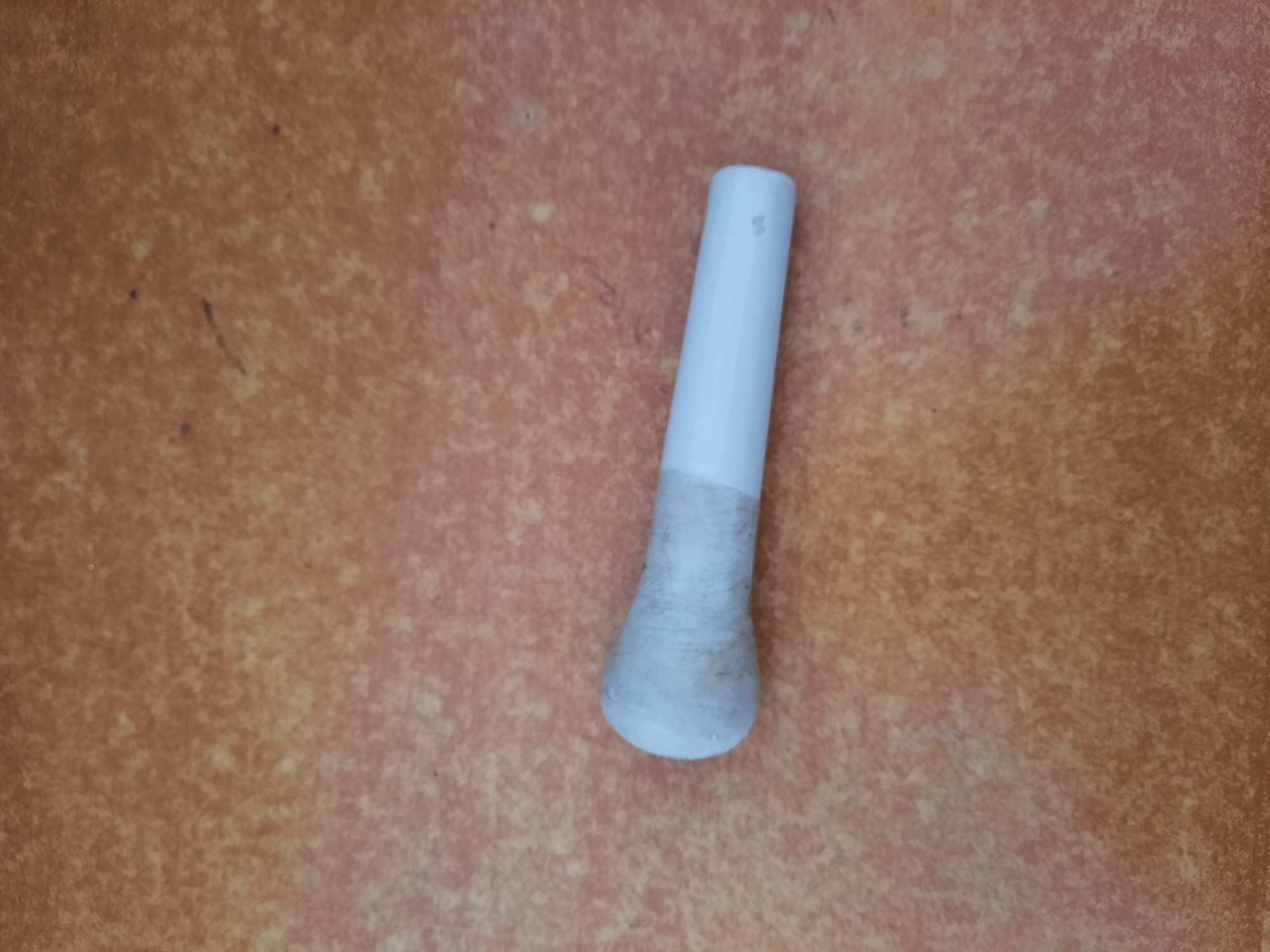 A white, cylindrical object with a rounded end is lying horizontally on a speckled reddish-brown surface. The rounded end is covered with a light-colored tape, which appears somewhat worn and stained. The rest of the object is smooth and white. The background surface has a texture resembling stone or tile, with variations in color and small speckles.