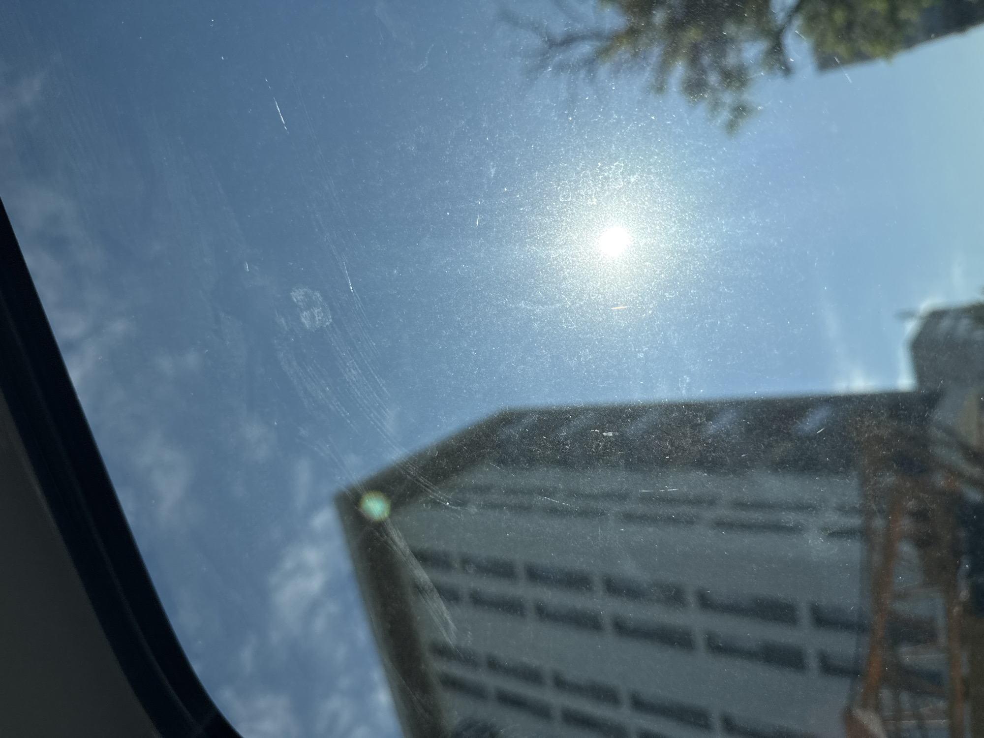 The image captures a view through a dirty window, likely from inside a vehicle, on a sunny day in Varna, Bulgaria. The sky is a clear blue with some white clouds. In the foreground, the window is streaked and smudged, with visible scratches.
On the left side of the image, a tall, modern building with a facade of repeating dark rectangular windows is partially visible. The building appears to be light-colored. A green glint, possibly a reflection or a light fixture, is present on the top edge of this building.
Towards the center and right of the image, the sun is a bright, glinting orb, casting a strong light that causes lens flare and washes out details in that area. A bare tree branch, silhouetted against the sky, is visible to the right of the sun. In the lower right corner, the top of another building, possibly a residential or office block, is visible, partially obscured by trees. In the foreground, at the bottom of the image, part of a structure with horizontal elements, possibly a balcony or a lower building section, is visible.
The overall impression is an urban landscape viewed through a less-than-pristine window, with the bright sunlight creating a somewhat hazy and glinting effect. The specific location in Varna cannot be identified definitively from the visual elements alone, beyond it being an urban environment.