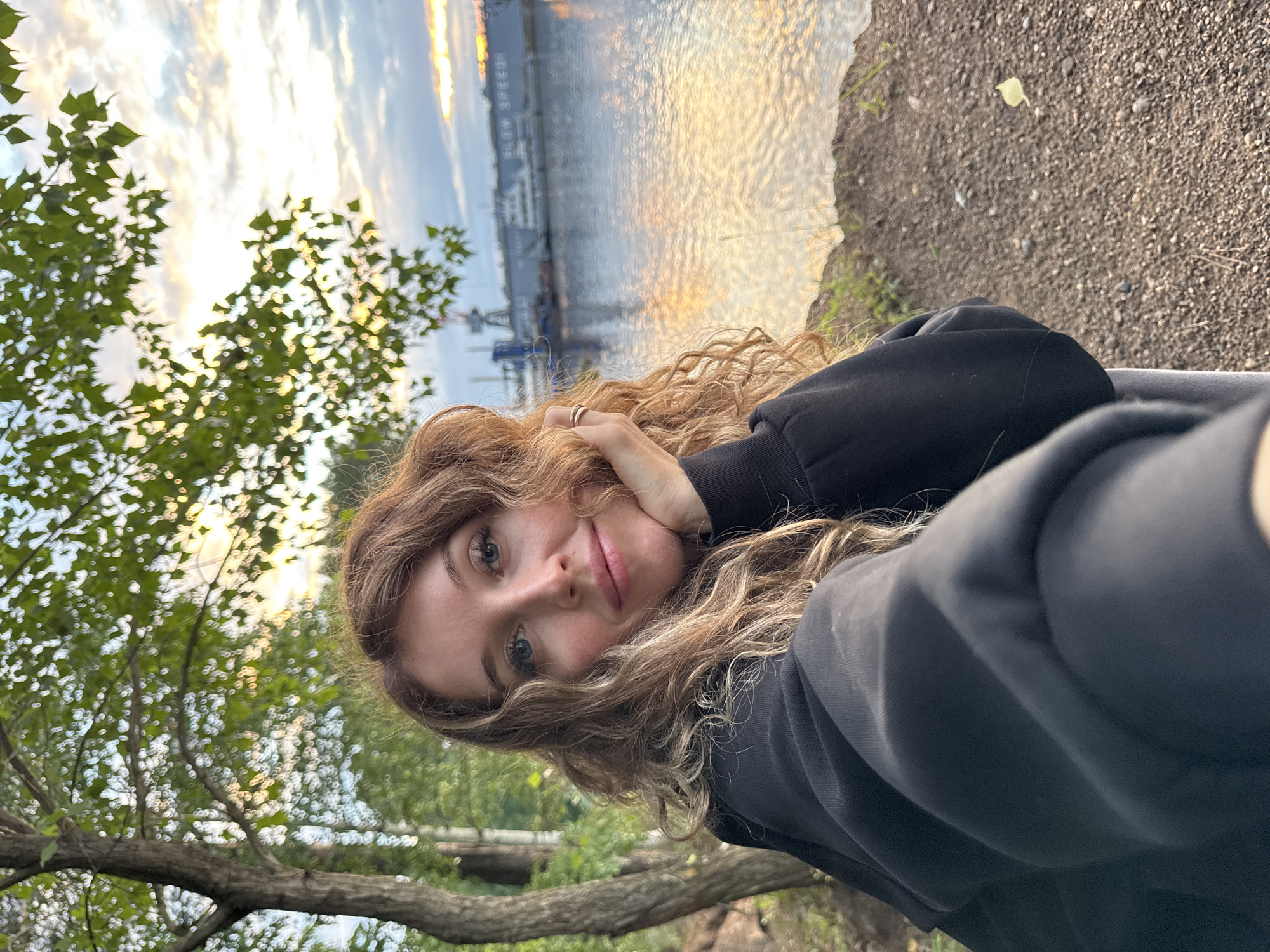 A young woman with long, wavy blonde hair is taking a selfie outdoors. She is wearing a black hooded sweatshirt. Her right hand is resting on her chin and she is looking directly at the camera with a slight smile. In the background, there is a body of water reflecting the sunset sky. On the far bank of the water, there is a large industrial structure with cranes. Trees and foliage are visible to the left of the woman. The ground in the foreground is covered with pebbles and dry leaves. This scene is in the city of Chaikovsky, Russia.