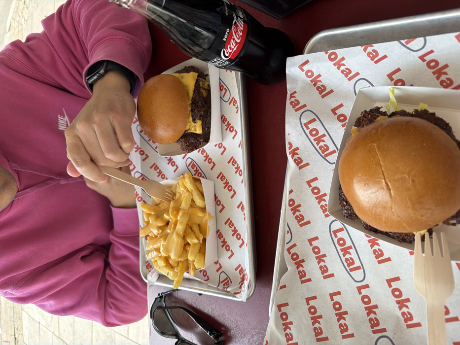 The image depicts a casual dining scene for two individuals at a Lokali Burgers restaurant in Tel-Aviv, Israel.
In the upper half of the frame, one individual is partially visible, wearing a hot pink hooded sweatshirt with a white "Nike" logo on the chest. Their right hand, adorned with a dark smartwatch, holds a light wooden fork poised over a white paper boat filled with golden-brown French fries smothered in orange-yellow cheese sauce. This meal is presented on a rectangular silver metal tray. Next to the fries, on the same tray, is a smash burger in another white paper boat, featuring a glossy bun, two beef patties, and melted yellow cheese. To the right of this tray, a dark green glass bottle of Coca-Cola Zero with a red label stands on the table. A pair of black sunglasses rests on the red table surface to the left of the tray.
In the lower half of the frame, a second, partially visible silver metal tray holds another meal. This includes a white paper boat containing a smash burger with a light brown bun and visible green lettuce. A second light wooden fork is placed next to this burger.
The table surface is a uniform deep red. Both metal trays are lined with white paper imprinted with a repeating pattern of the "Lokali" brand logo in black text within an oval, and smaller "Lokali" text, confirming the establishment's identity. The background is out of focus, indicating an indoor or outdoor dining setting.