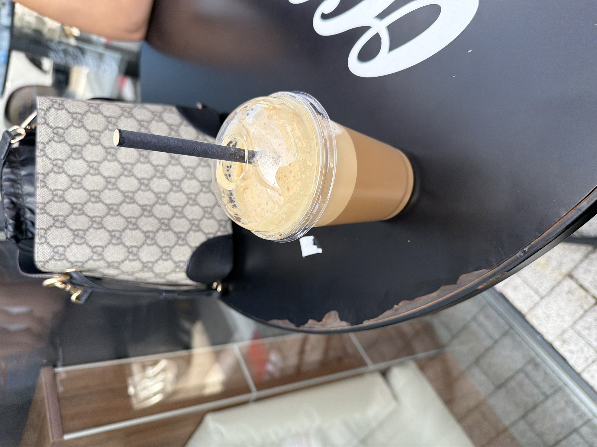 In Chuchuligovo, Bulgaria, on a black, circular table with a distressed edge, sits a clear plastic cup filled with a frothy, light brown beverage, topped with a domed lid. A dark gray paper straw extends from the center of the lid. Behind the cup, a beige handbag with a repeating "GG" logo pattern and black trim is visible. The background shows a reflective surface that appears to be a glass window, with a wooden structure to the left and hints of a car and building beyond. White lettering, partially obscured, is present on the table to the right.