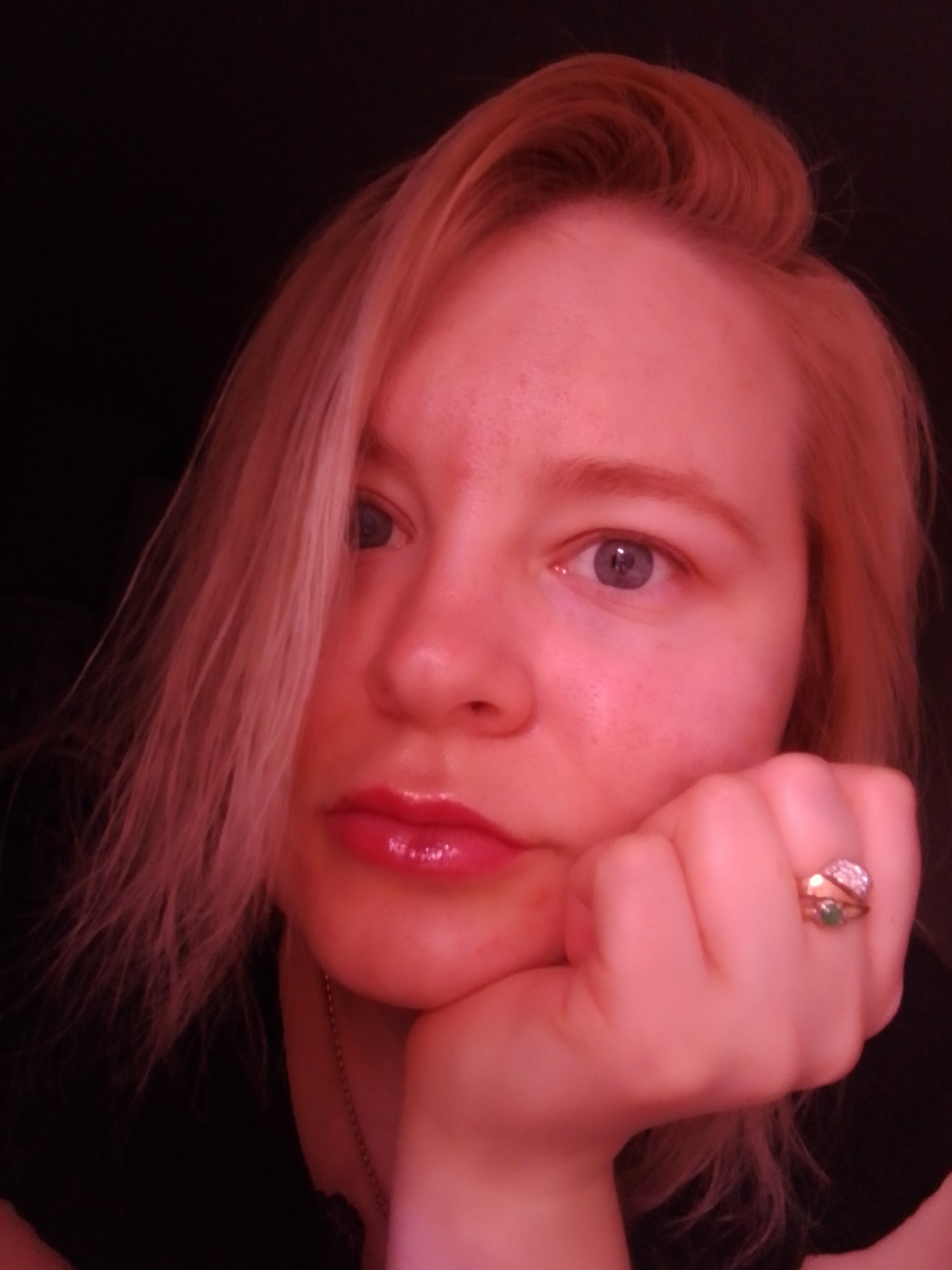 A close-up, selfie-style photograph shows a fair-skinned individual with light blonde hair, resting their face in their hand. The hair is parted to the side, with some strands falling across their face. The person has light eyes and glossy lips. A ring is visible on the hand supporting the face. The background is dark, suggesting a dimly lit indoor setting. The photograph exhibits a reddish hue, possibly due to the lighting conditions or a filter applied to the image. There is no indication of the image being taken in Izhevsk, Russia.