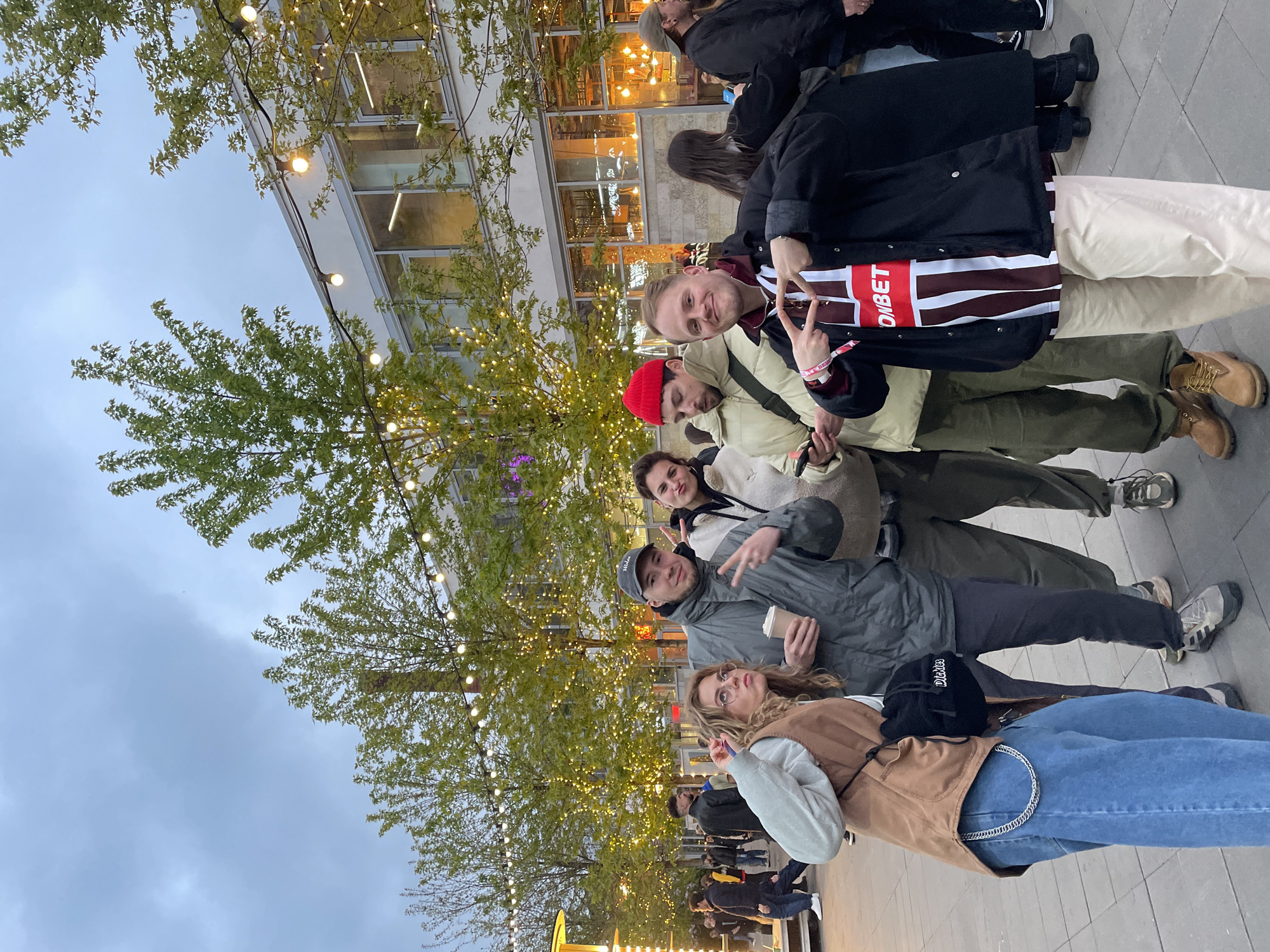 A group of six people is posing for a photograph in an outdoor urban setting. They are standing on a paved area that appears to be a pedestrian zone. In the background, there is a building with large windows and what looks like a cafe or restaurant inside, illuminated by warm lights. Above and around them are trees decorated with string lights, suggesting a festive or decorative atmosphere. The sky is overcast.
The people in the foreground are dressed in casual clothing, including jackets, jeans, and various accessories like hats and scarves. Some are making peace signs with their fingers. The image quality is clear, allowing for the visibility of details like facial expressions and clothing textures.
Based on the clothing and background, it is impossible to confirm this image was taken in Saint Petersburg, Russia.