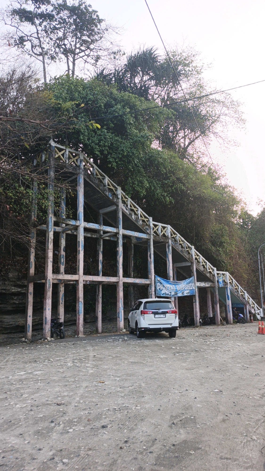 A white SUV is parked on a gravelly surface in front of a tiered, concrete structure that ascends a hillside covered in dense green foliage. The structure has a faded, weathered appearance with peeling paint in shades of blue and pink on its pillars and railings, which are adorned with yellow accents. A blue banner with white lettering, reading "PANTAI BATUHIIU", is suspended from the structure. A motorcycle is parked to the left of the SUV. In the background, trees and vegetation cover the hillside. The sky above is a pale, hazy pinkish-white. Power lines are visible stretching across the upper portion of the frame. The scene appears to be an outdoor location in Desa Ciliang, Indonesia, likely a pathway or viewing area leading to a beach.