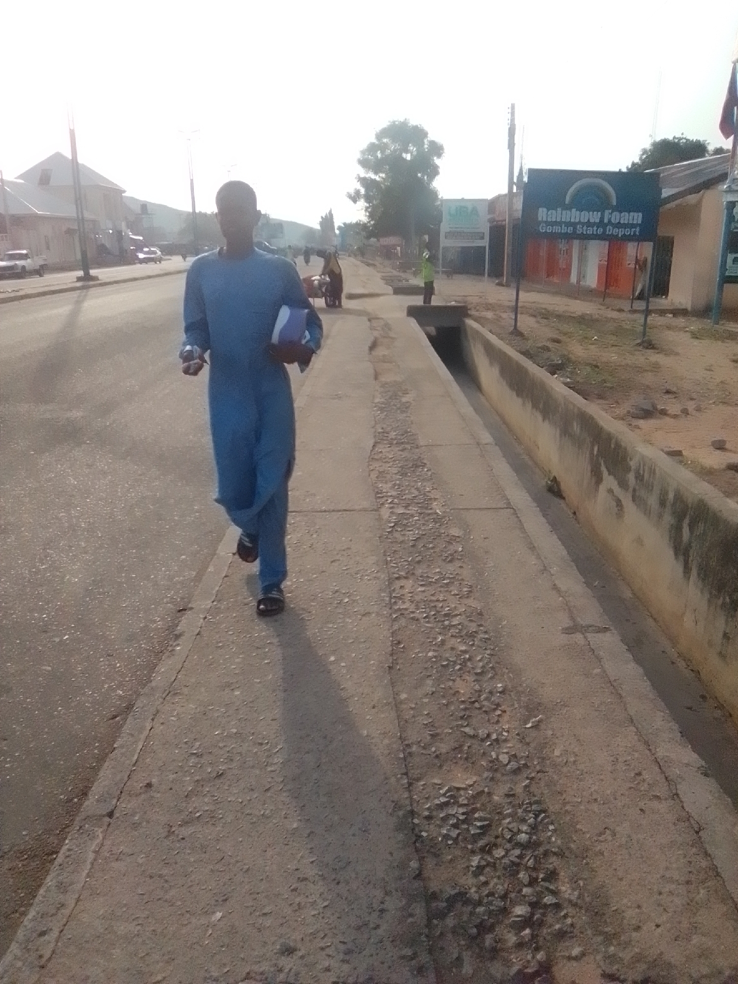 The image depicts a person wearing a blue robe and sandals, walking on a sidewalk. The person is carrying a blue and white object in their left hand. The sidewalk is made of concrete and appears to be in a state of disrepair, with visible cracks and loose stones. To the right of the sidewalk is a concrete drainage channel. In the background, there is a street with vehicles, buildings, and trees. A sign with "Rainbow Foam Gombe State Deport" is visible. The scene is outdoors during the day, with bright sunlight casting shadows. The overall environment suggests an urban setting in Pantame, Nigeria, with elements of everyday life and infrastructure.