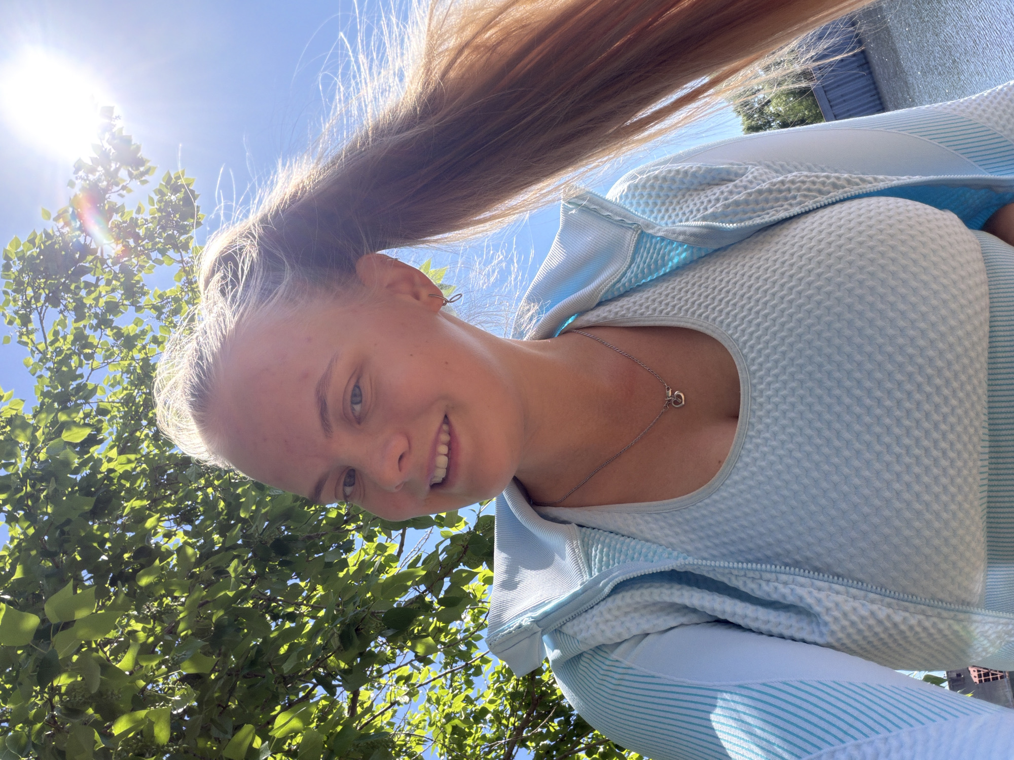 A smiling, fair-skinned young woman with blond hair in a ponytail is pictured in a selfie-style image. She is wearing a light blue textured top and a light blue zip-up hoodie. A silver necklace with a heart-shaped pendant is visible. The background features green foliage from a tree and a bright blue sky with the sun shining. The exact location in Chaikovsky, Russia cannot be determined from the image alone.