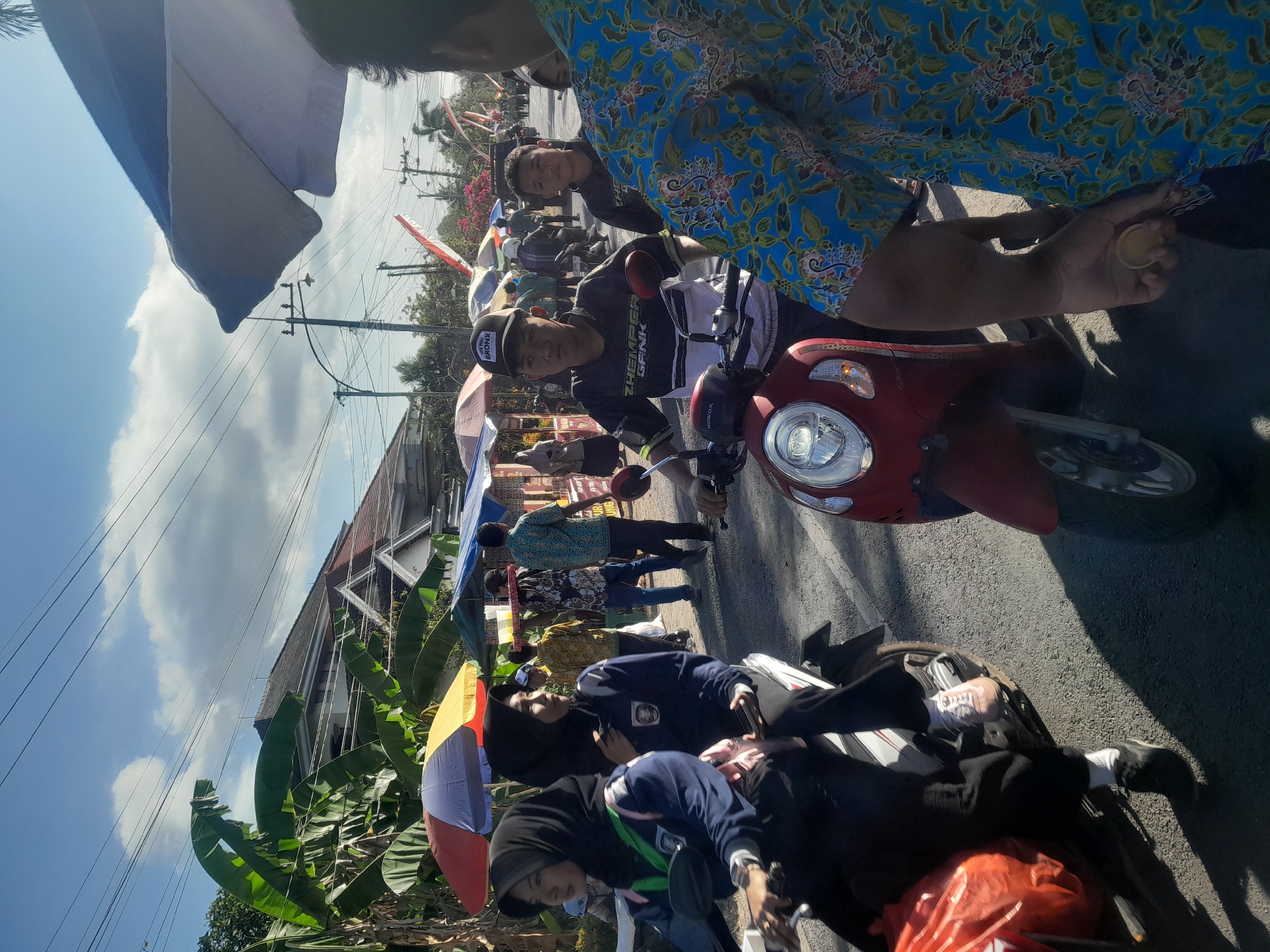 In Sambirejo, Indonesia, a street scene unfolds with people on motorcycles under a bright, cloudy sky. A woman in a hijab rides a white scooter on the left, a large orange bag on her lap. Beside her, another woman in a hijab also sits on a scooter. In the center, a young man in a black cap and a patterned shirt rides a red scooter, looking forward. Behind him, another young man on a motorcycle also looks ahead. The street is lined with market stalls with colorful umbrellas, and a building with a tiled roof is visible in the background. Power lines crisscross the sky. The overall atmosphere suggests a busy, outdoor gathering or market event.