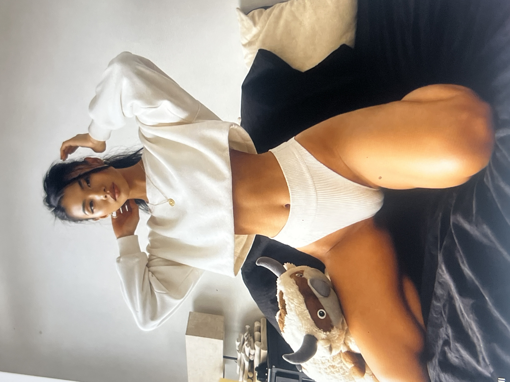 A young Asian woman with dark hair is posing on a bed in a room with white walls. She is wearing a white crop top and white underwear. She has her hands on her head. A stuffed animal, resembling a bison, is resting on her lap. The room appears to be a bedroom, with pillows and bedding visible.