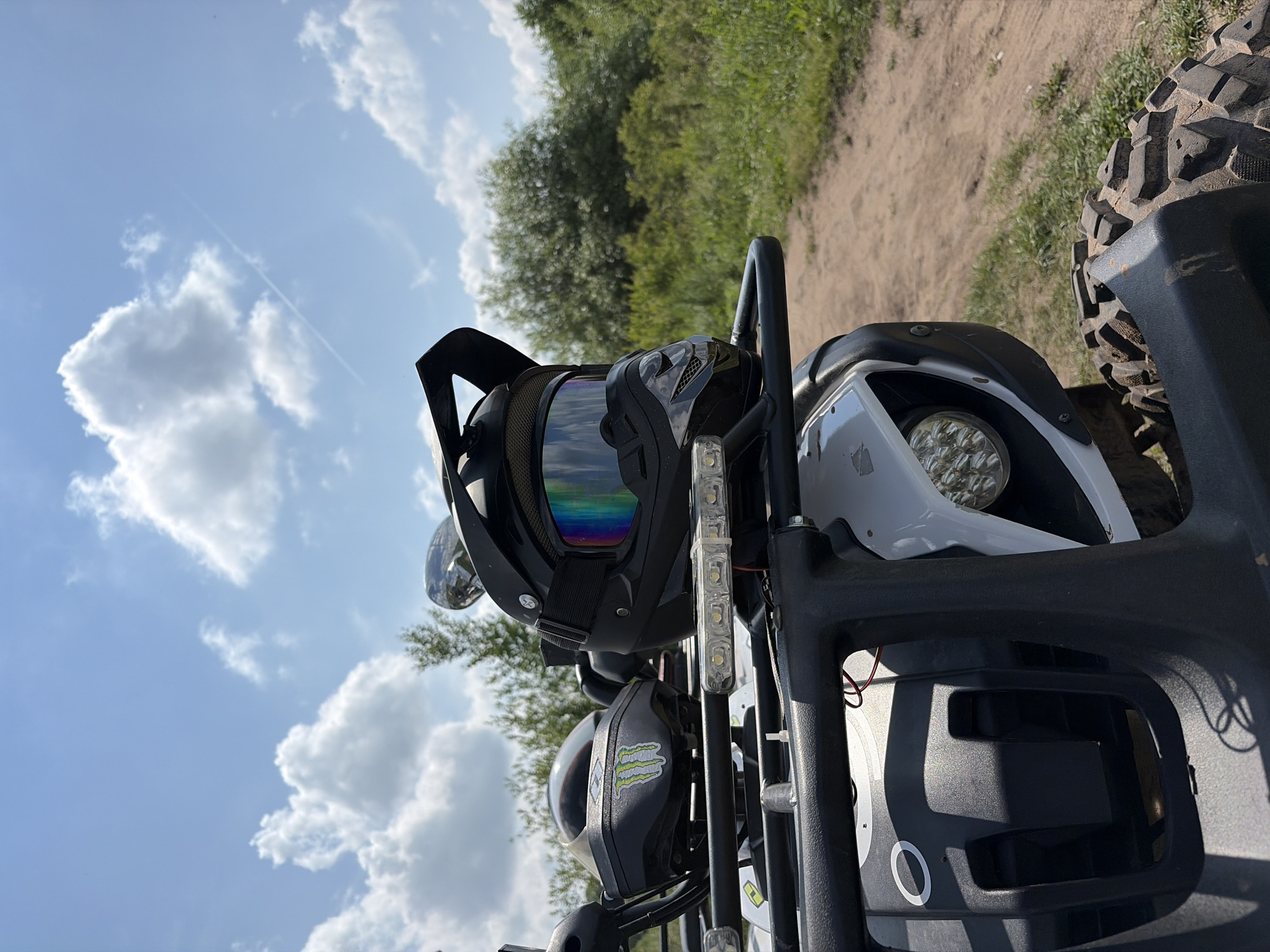 This is a close-up, low-angle shot of a black helmet with rainbow-tinted goggles resting on the front rack of a white ATV. The ATV is parked on a dirt path that curves to the right, bordered by green grass and dense foliage. In the background, the sky is a bright blue with fluffy white clouds and a faint contrail from an aircraft. Another helmet, this one white with a green "Monster Energy" logo, is visible behind the black helmet on the ATV's rack. The ATV's headlight is also visible, along with part of its tire. The scene appears to be set in a rural or outdoor recreational area, possibly a trail or park. The image is taken in Gushchino, Russia.