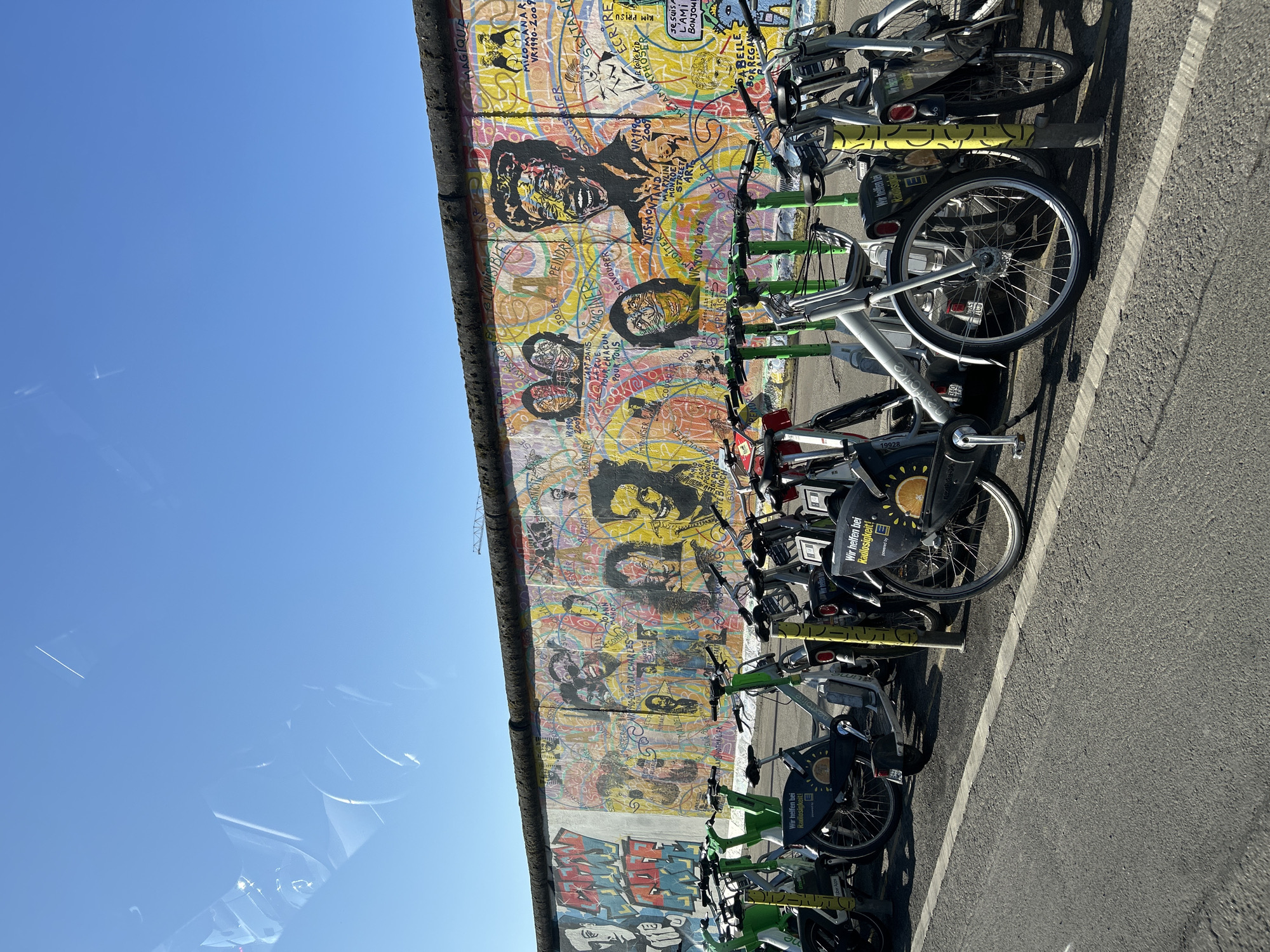 The image presents a vibrant outdoor scene in Berlin, Germany, capturing a segment of the iconic Berlin Wall, extensively covered in colorful street art. A long line of shared bicycles is neatly parked along the base of the wall on an asphalt ground, marked by a white line.
The wall itself is a canvas of intricate graffiti and murals, featuring various styles, abstract patterns, and detailed portraits. Recognizable figures within the artwork include stylized depictions of Marilyn Monroe and Ray Charles, accompanied by text such as "MARYLIN MONROE" and "1990-2009 RAY CHARLES". Other phrases and words are scattered across the wall, including "MAGIQUE," "SEXTASIF," "ÉCRIRE," "L'ART DANS POUR TOUS," "STREET ART," and "IMPROVISER SCULPTER," reflecting a rich tapestry of urban expression.
The foreground is dominated by a collection of rental bicycles, likely from services like Nextbike and Lime, distinguished by their silver, green, and yellow frames. Visible text on the bikes includes "Uber Apps," "Wir helfen bei Radlosigkeit!" (We help with bike-lessness!), "powered by E," and bike ID "19928". These bikes are parked on a grey asphalt surface, where prominent shadows cast by the bikes indicate bright sunshine.
The sky above is a clear, brilliant blue, suggesting a sunny day, likely mid-day or early afternoon. Faint reflections are visible in the upper left of the frame, possibly from a window. The scene captures the blend of historical significance, contemporary street art, and modern urban mobility in Berlin.