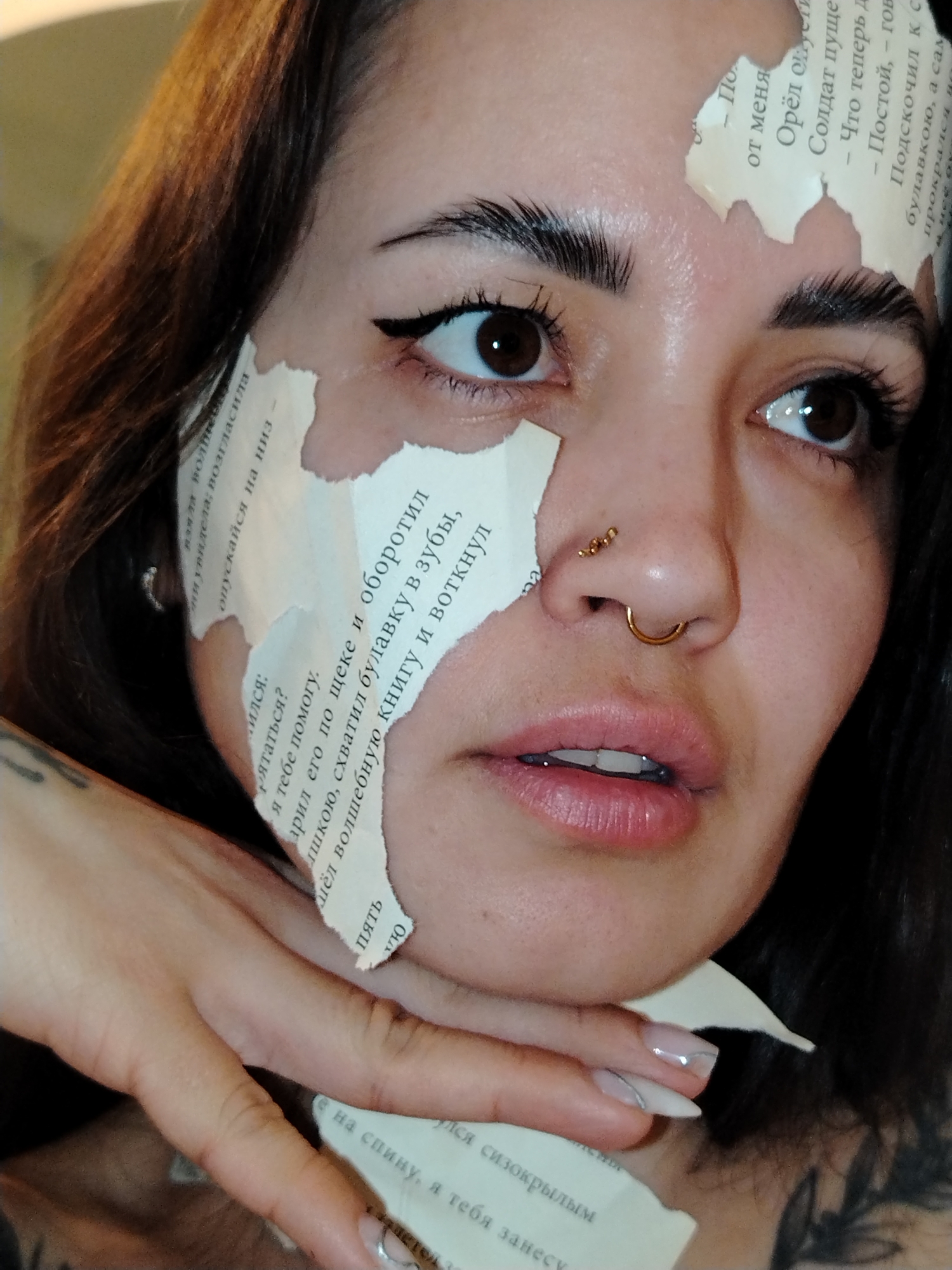 A close-up shot shows a person with fair skin, dark brown hair, and dark eyes. They are wearing nose piercings—a small stud near the top of their nostril and a gold hoop through their septum. Their makeup includes eyeliner and defined eyebrows. Pieces of paper with Russian text are adhered to their face, covering parts of their forehead, cheek, and the side of their nose. The person's hand, with pale nails adorned with a silver design, is positioned near their chin. Tattoos are visible on their hand and neck. The environment is blurred, suggesting an indoor setting with warm lighting. It is not possible to determine that this image was taken in Noviy, Russia.