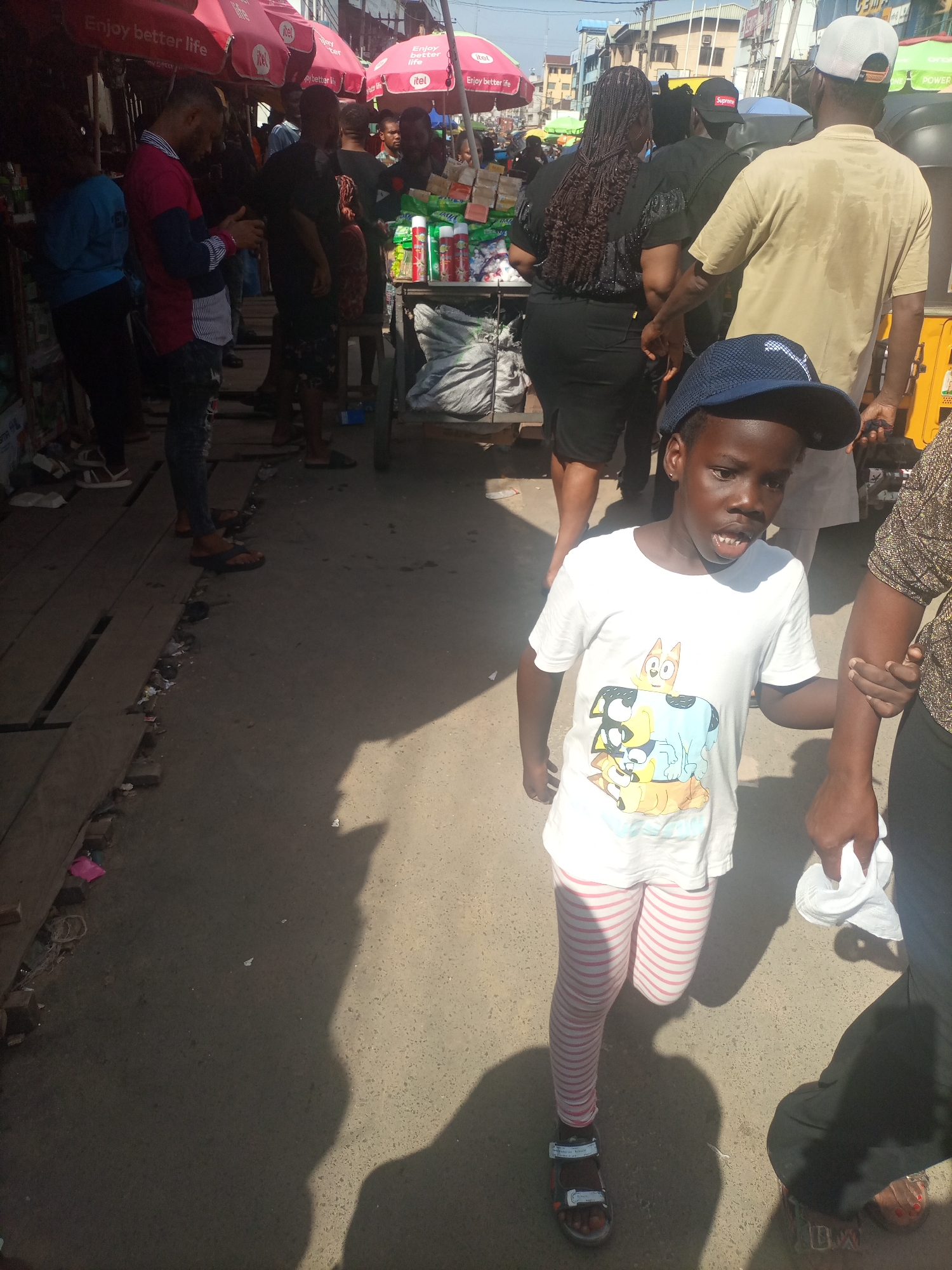 This media file captures a bustling street scene in Onitsha, Nigeria, likely during the daytime given the strong shadows. The foreground is dominated by a young child wearing a white t-shirt featuring cartoon characters and striped pink and white leggings. The child is wearing sandals and a blue baseball cap, and appears to be walking or running, holding onto the arm of an adult whose legs and hand are partially visible on the right side of the frame.
The background reveals a lively market environment. Several adults are present, some browsing at stalls, others walking. Umbrellas, likely for shade, are visible, some displaying text such as "Enjoy better life" and "itel." Stalls are laden with various goods, including packaged items and what appear to be sacks of produce or goods. The ground is a mixture of concrete and wooden planks, suggesting a well-trodden path within the market. The overall impression is one of vibrant commerce and daily life. The presence of people in diverse attire and the nature of the market stalls point to a busy urban setting.