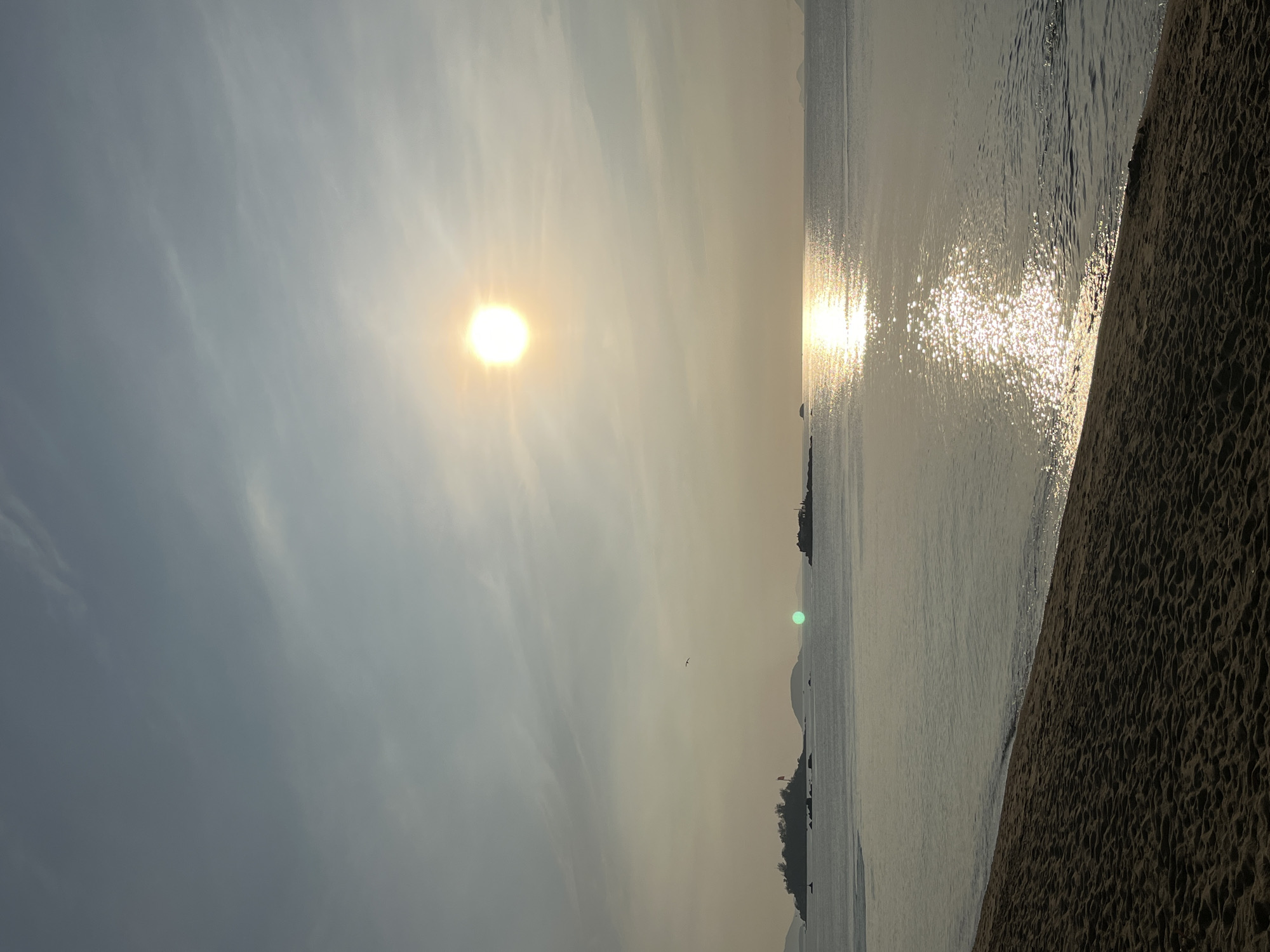 The image shows a coastal scene, likely a beach, in Nha Trang Ward, Vietnam. The sun is visible in the sky, with its reflection shimmering on the water's surface. Sand covers the foreground, indicating the beach. There are distant land masses in the sea and a bird flying in the sky.