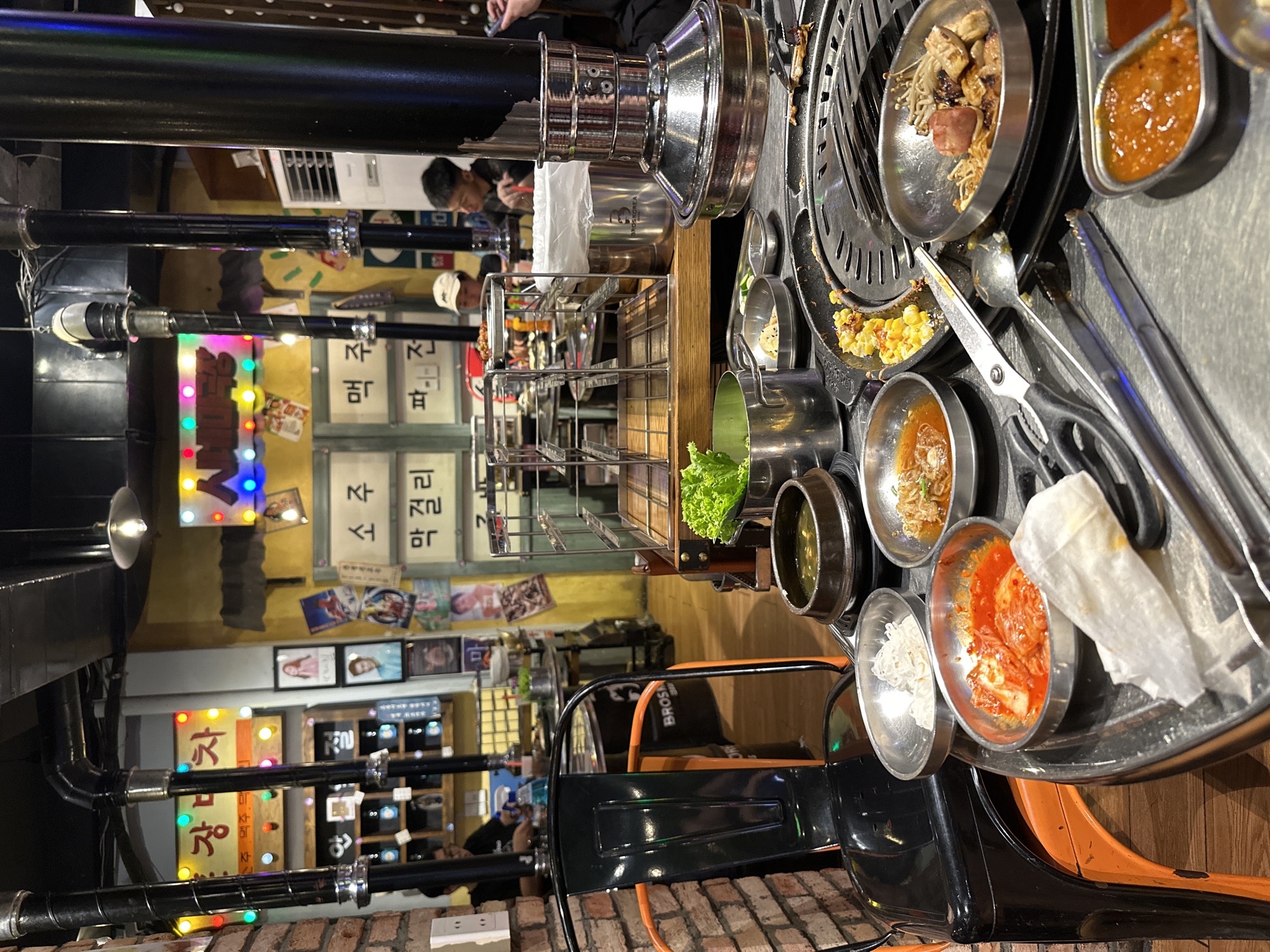 This indoor photo depicts a Korean BBQ dining setup in Thủ Đức, Vietnam. The scene is captured at an angle from the perspective of someone seated at the table. The main focal point is a circular, stainless steel Korean BBQ grill positioned in the center of the table. Around the grill, there are various small metal bowls containing different Korean side dishes such as kimchi, corn, salad, sauces, and mushrooms with grilled meat. Utensils such as spoons, tongs, and scissors are also visible near the grill.
In the background, there are other people dining at nearby tables. The decor includes a wall with Korean lettering and lighted signs. There are exposed black pipes and light fixtures near the ceiling. The table is set with a black and orange chair. The overall lighting appears to be artificial and somewhat dim, creating a casual dining atmosphere.