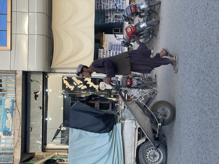 The image displays a daytime street scene in Kandahar, Afghanistan. A man, dressed in a dark purple shalwar kameez, an olive-green waistcoat, and a patterned pakol-style hat, walks from left to right across a paved street, pushing a two-wheeled metal handcart. His feet are clad in dark sandals. The handcart features a small front wheel and a white net attached to its front frame. To the immediate left, a portion of a light-colored utility truck is visible, with its bed covered by a light blue tarp and an additional black tarp.
In the background, on the right side of the street, three motorcycles are parked, including a red one with a rear plate-like element displaying "20" and a stylized "R". The street is lined with commercial buildings. The building on the left features a ground-level glass storefront that is internally lit, revealing what appears to be a light-colored mannequin. Above this, a grey panel is present, topped by a blue-railed balcony with a sign depicting text in an Afghan script and images of clothing. The building on the right has a modern grey paneled upper facade, while its ground-level storefront is partially obscured by a large white fabric covering. Stacks of white boxes, some labeled "MILAD," are visible behind this covering. The sky overhead is clear blue.