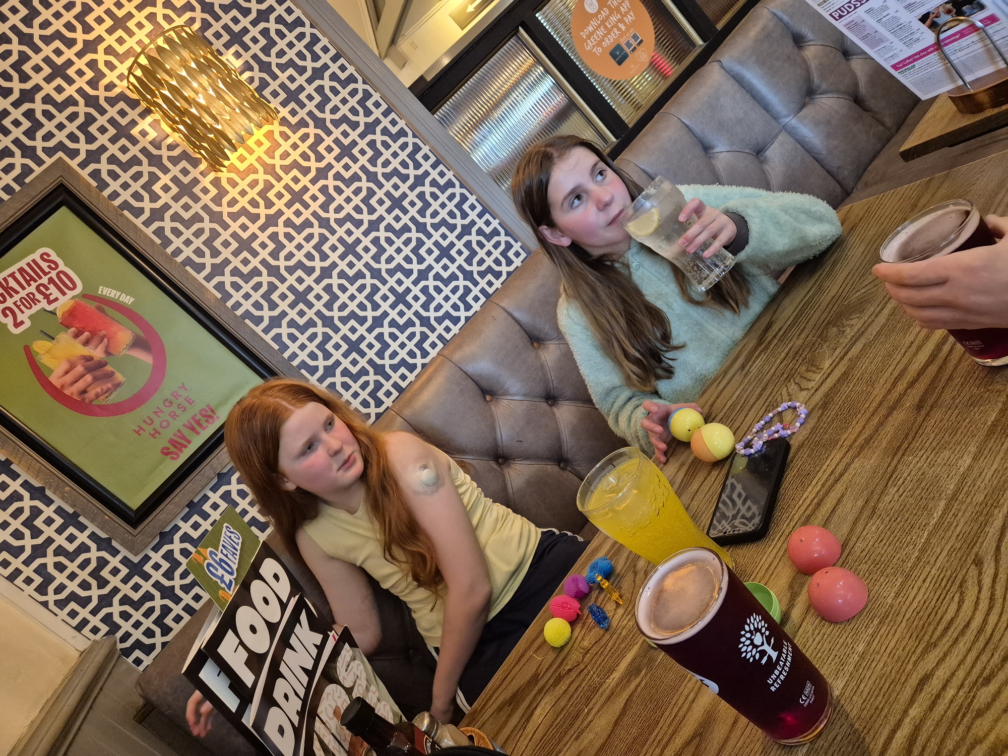 The image shows two young girls sitting at a table in what appears to be a pub or restaurant. The girl on the left has red hair and is wearing a yellow top and black shorts. She has a medical device attached to her arm. The girl on the right has brown hair and is wearing a light green sweater, and is drinking from a glass with lemon slices.
On the table, there are several objects including a smartphone, toy balls, a purple bead bracelet, and drinks in pint glasses. One drink is golden in color, the other is dark.
The background shows a padded booth seating area and patterned wallpaper with a framed advertisement for cocktails. There are also menus on the table. The OCR text includes "HUNGRY HORSE" which is a chain of pubs in the United Kingdom. This image was probably taken at a Hungry Horse pub. The OCR also has "GREENE KING APP" which is another chain of pubs in the United Kingdom. The image might be taken at a pub from either the Hungry Horse or Greene King chains.