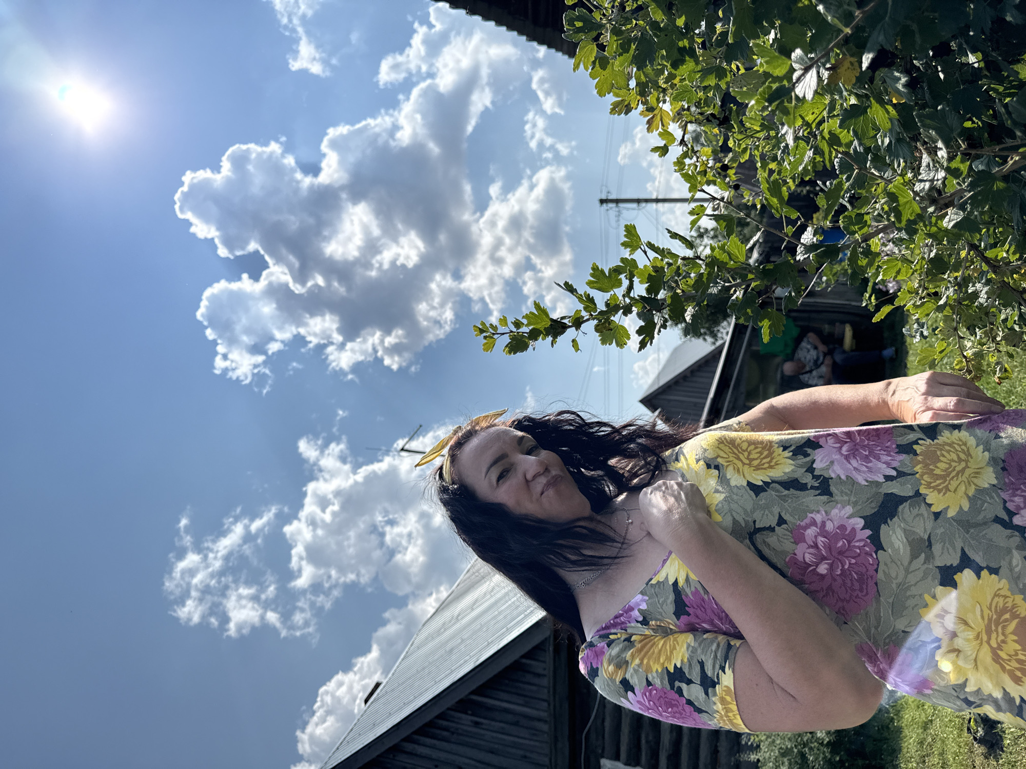 In Zasechnyi, Russia, a woman with dark hair, wearing a floral dress, poses in front of a wooden structure under a bright, sunny sky with fluffy white clouds. The woman is looking towards the camera with a slight smile and her hand on her chest. To the right of the woman, a leafy green bush is visible, and in the background, another building and a utility pole can be seen. The scene appears to be set in a rural or semi-rural outdoor environment.