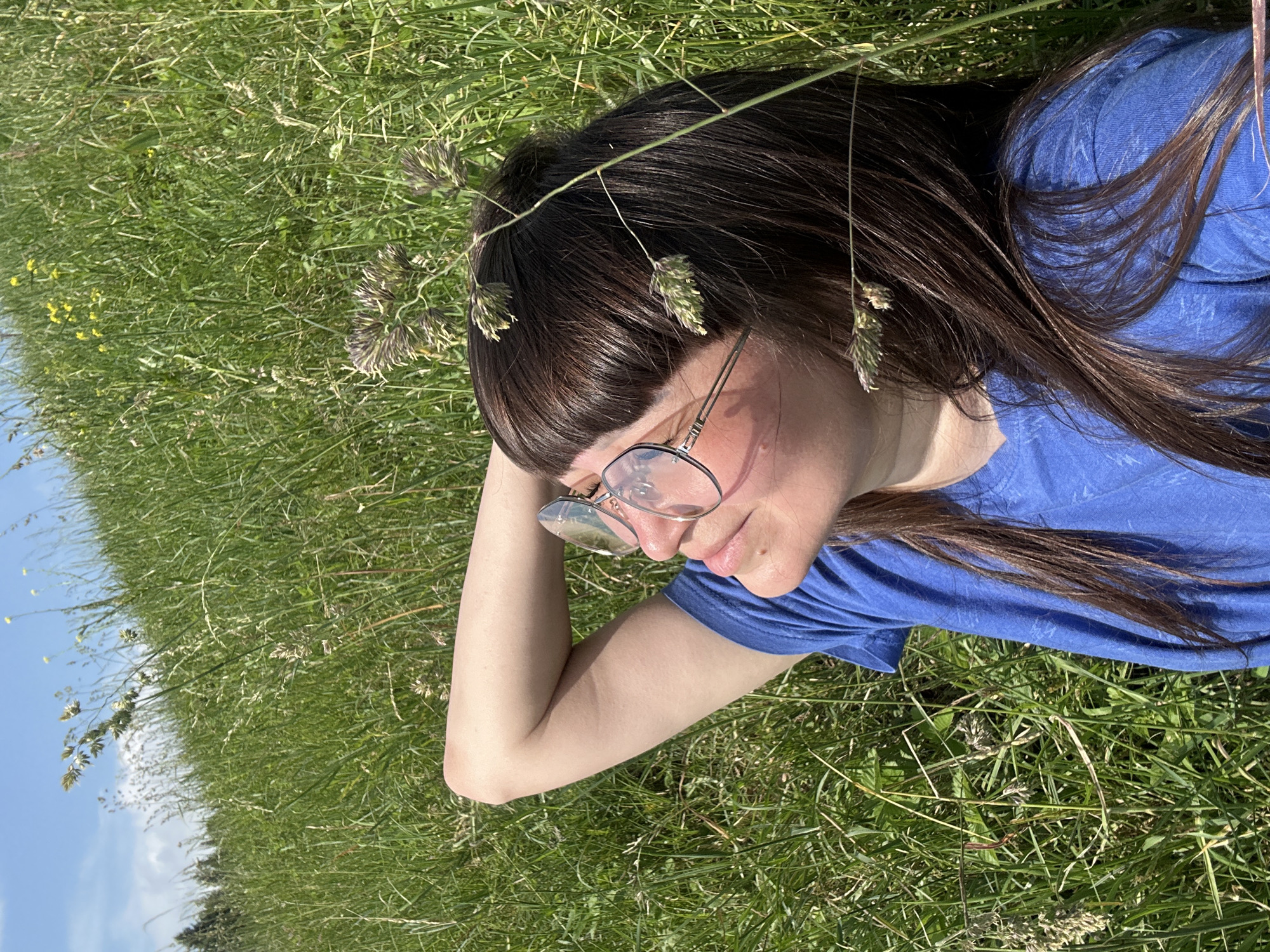 A young woman with dark brown hair and bangs, wearing a blue t-shirt and clear-rimmed glasses, lies in a grassy field. She is on her back, with her left arm bent and resting behind her head, looking upwards. Her face is turned to the right, and her eyes are closed, suggesting she is enjoying the sun. The field is lush and green, with tall grass and some small yellow flowers visible in the background. The sky is blue with some scattered white clouds. The overall impression is one of peaceful relaxation in nature. The image depicts a scene in a grassy field in the vicinity of Buranovo, Russia.