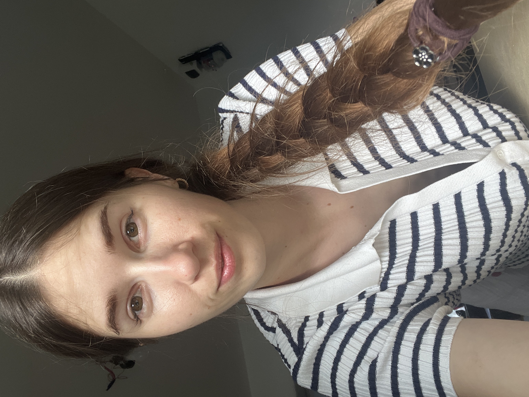 A young Caucasian woman with long, braided, reddish-brown hair is shown in a close-up, over-the-shoulder shot. She has fair skin with freckles and light brown eyes. She is wearing a white and navy striped collared shirt. The background is dimly lit and appears to be indoors, with a faint hint of what might be a window or a wall in the upper right corner. There is a dark object hanging from her hair on the left side of the frame.